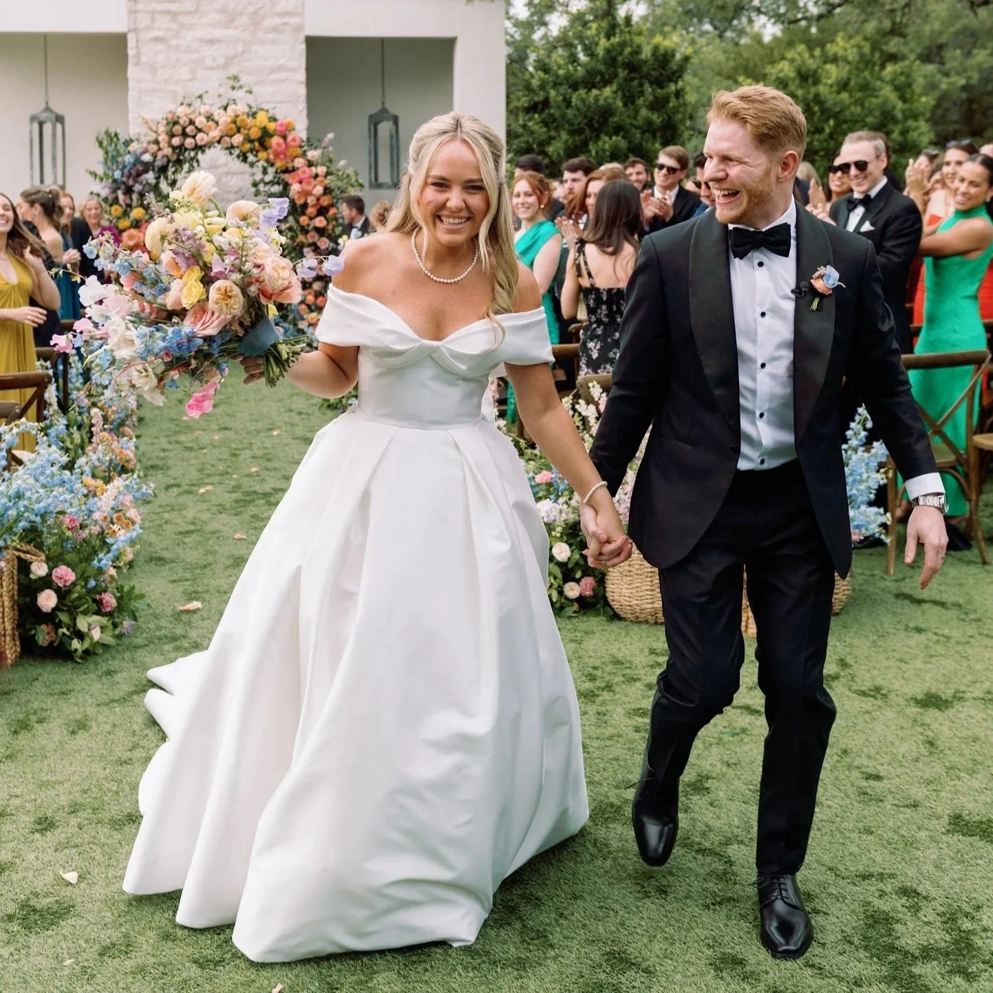 A bride and groom smiling and holding hands as they walk outdoors during their wedding celebration, with friends and family in the background, surrounded by flowers and greenery.