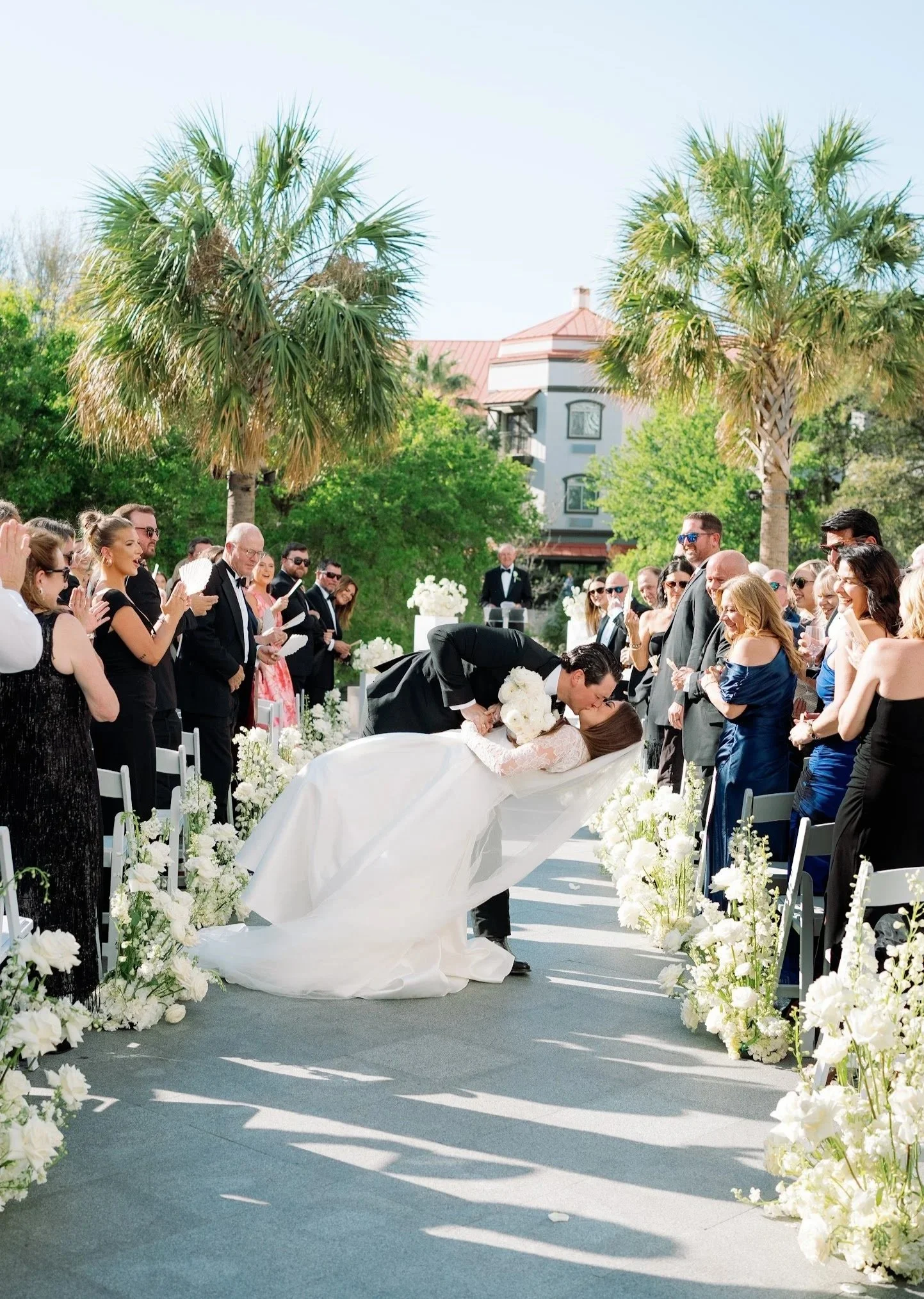 A wedding ceremony outdoors with a bride and groom sharing a kiss, surrounded by guests and floral decorations, in a sunny setting with palm trees.