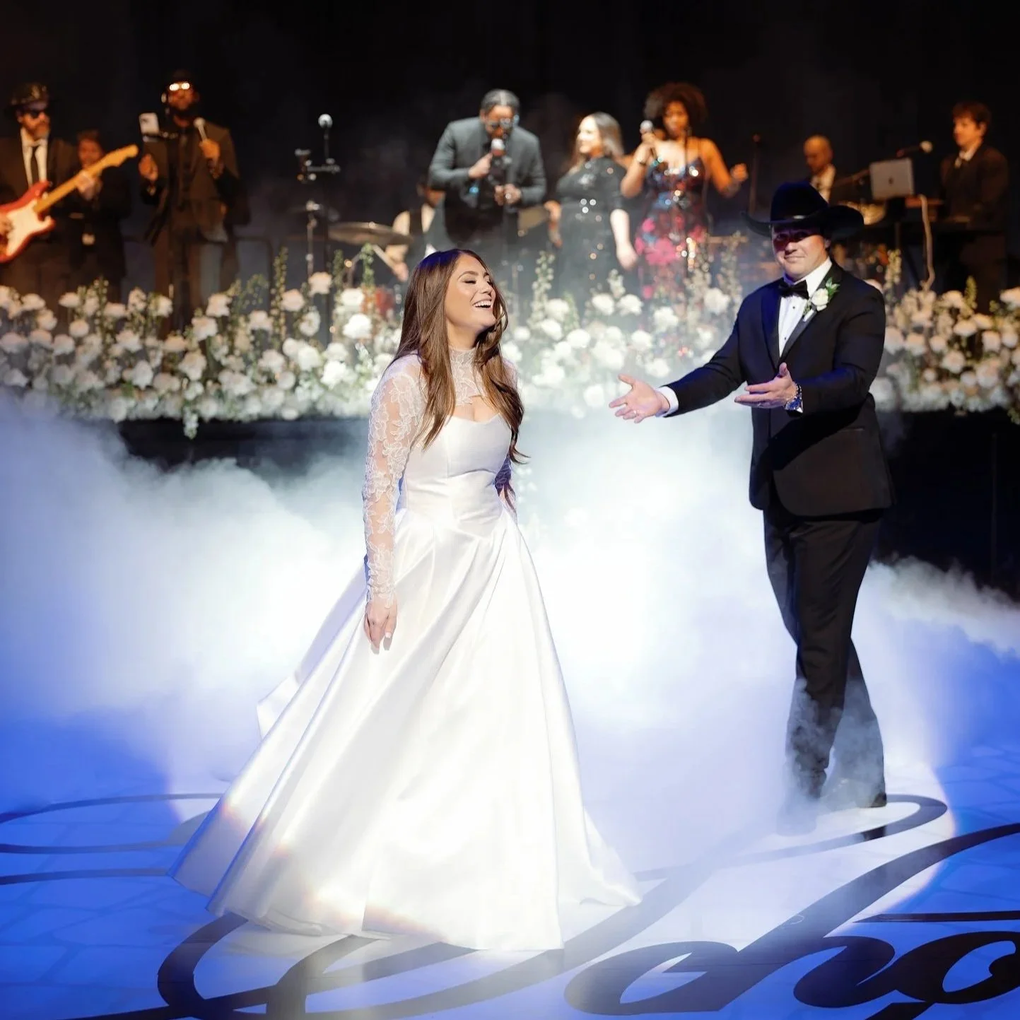 A bride in a white wedding gown and a groom in a black tuxedo with a cowboy hat dance on a foggy dance floor during a wedding reception, with a band playing live music in the background.
