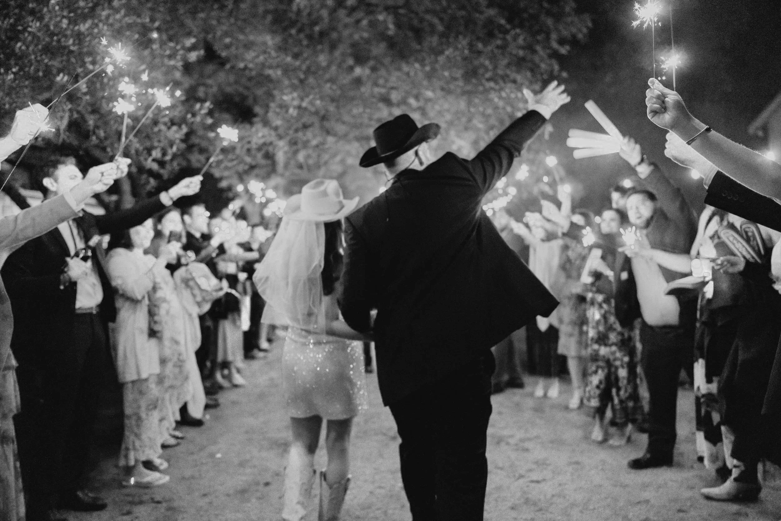 People celebrating at night with sparklers, some wearing traditional attire. One person in the center is wearing a hat and appears to be dancing. The crowd is closely gathered outdoors, with trees in the background.