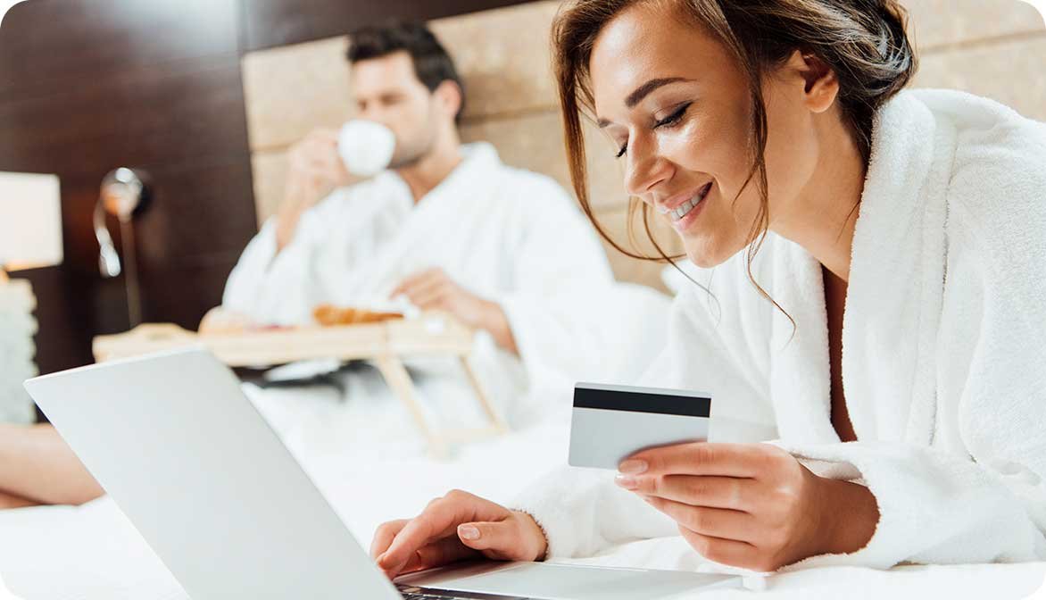 A woman sitting on a hotel bed using a laptop and credit card to make an online purchase, illustrating how guests redeem targeted coupons and promotional offers during their stay.