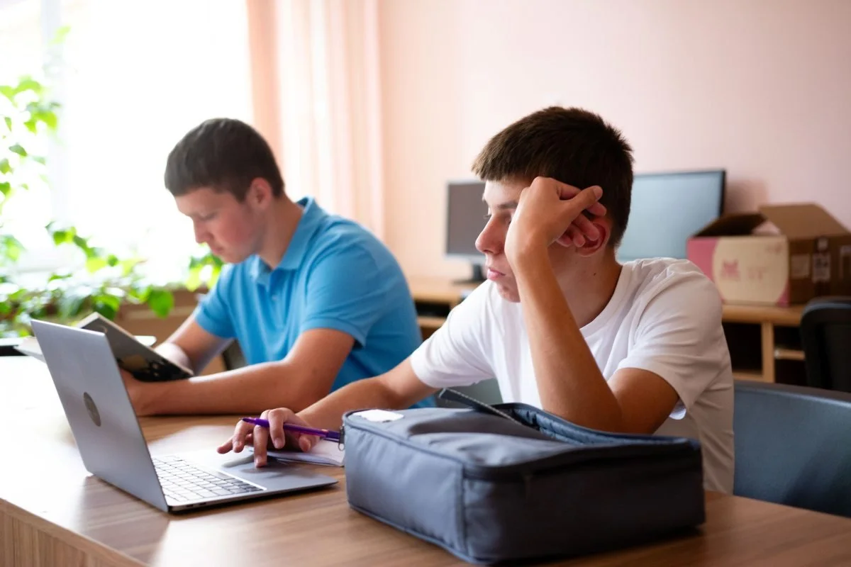 Teenagers studying at a table with a laptop and books. One student looks pensive while the other focuses on reading during their study session
