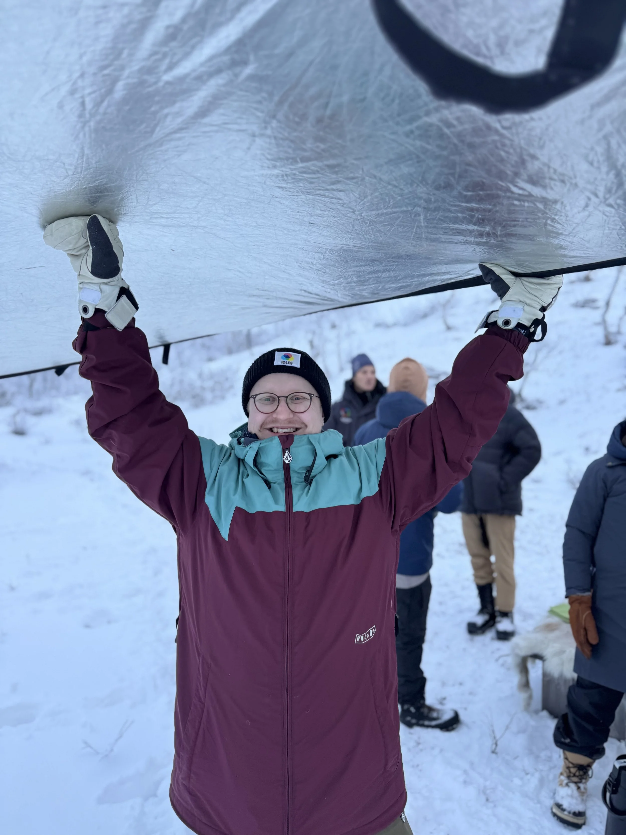 En man bär vinterskit och mössa, håller upp en stor spegel eller liknande föremål ovanför huvudet, utomhus i snö, med andra människor i bakgrunden.