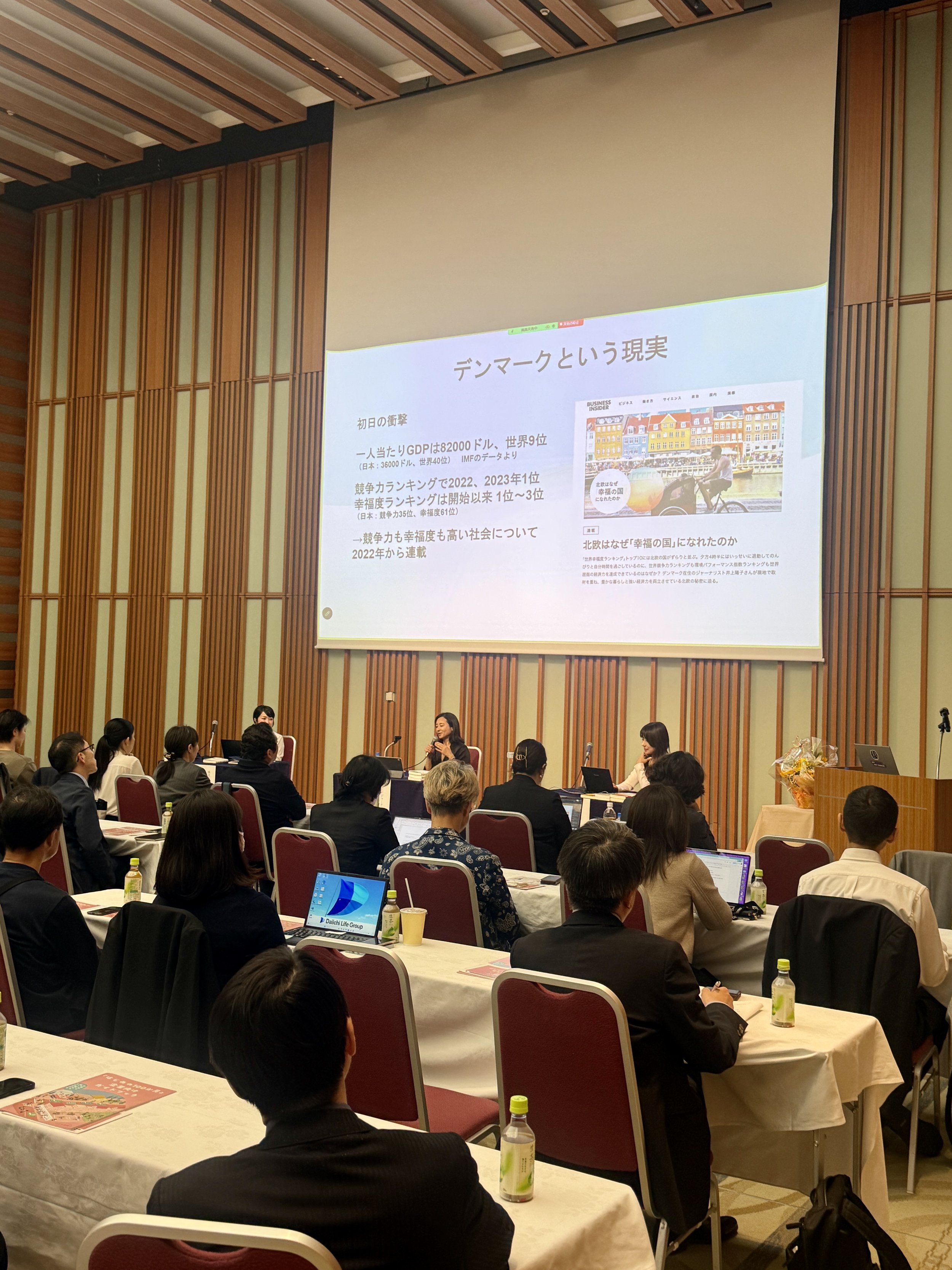 Yoko Inoue giving a talk at Keidanren Hall in Tokyo, a leading venue of Japan's business community, with Japanese text displayed on the screen behind her.