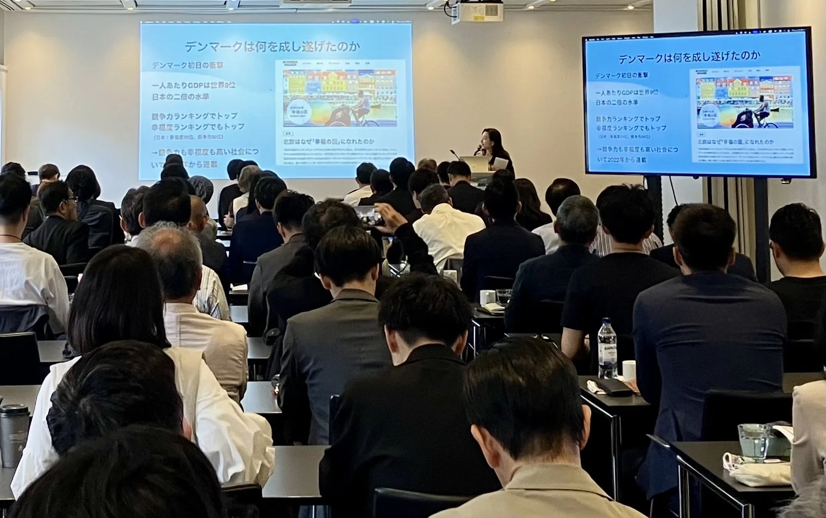 Yoko Inoue speaking at a podium to a large audience, with her presentation on Denmark projected on a screen behind her.