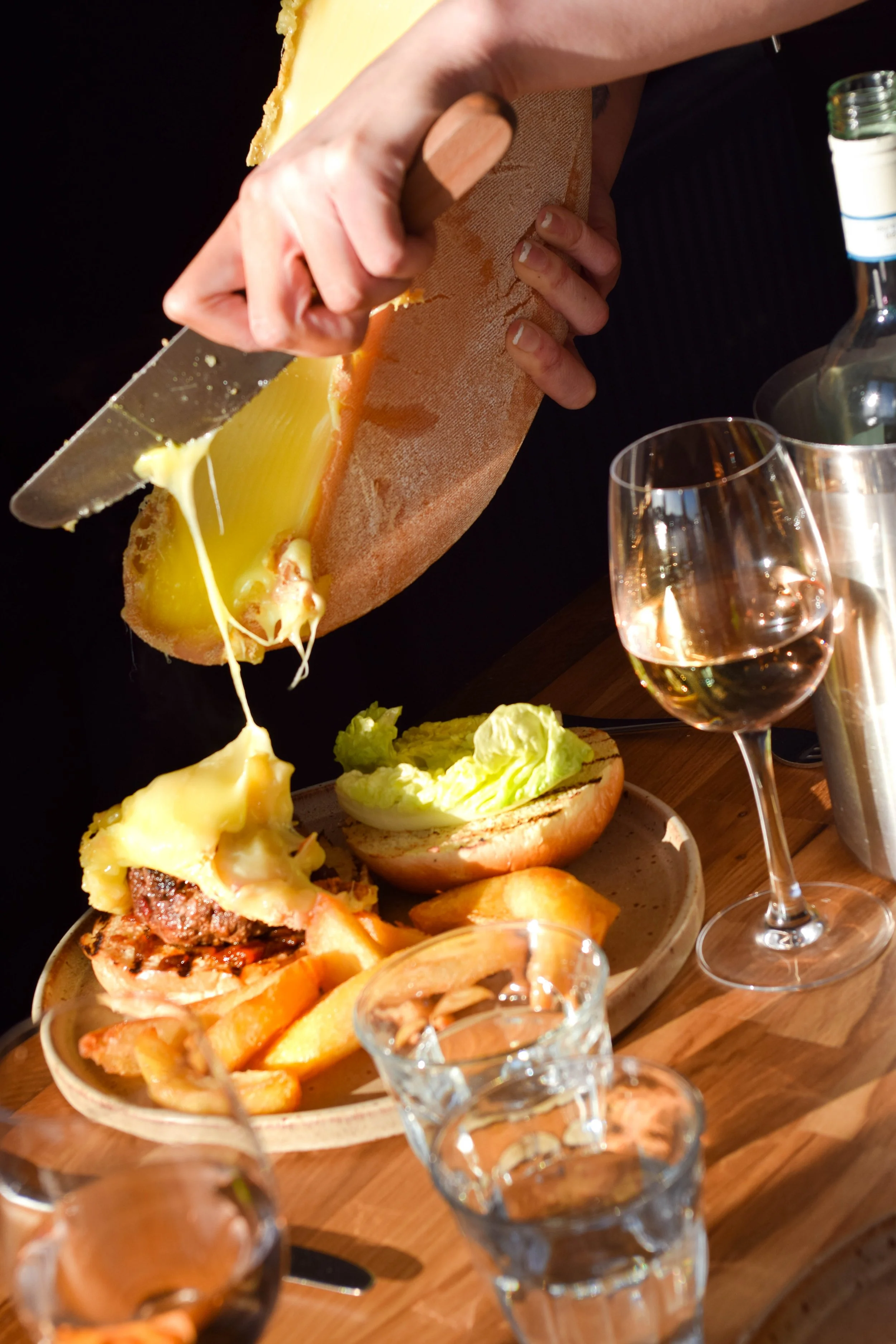 A person, slicing melted Raclette cheese over a plate with a burger, fries, lettuce, and a bread roll, with a glass of red wine and a bottle in the background.