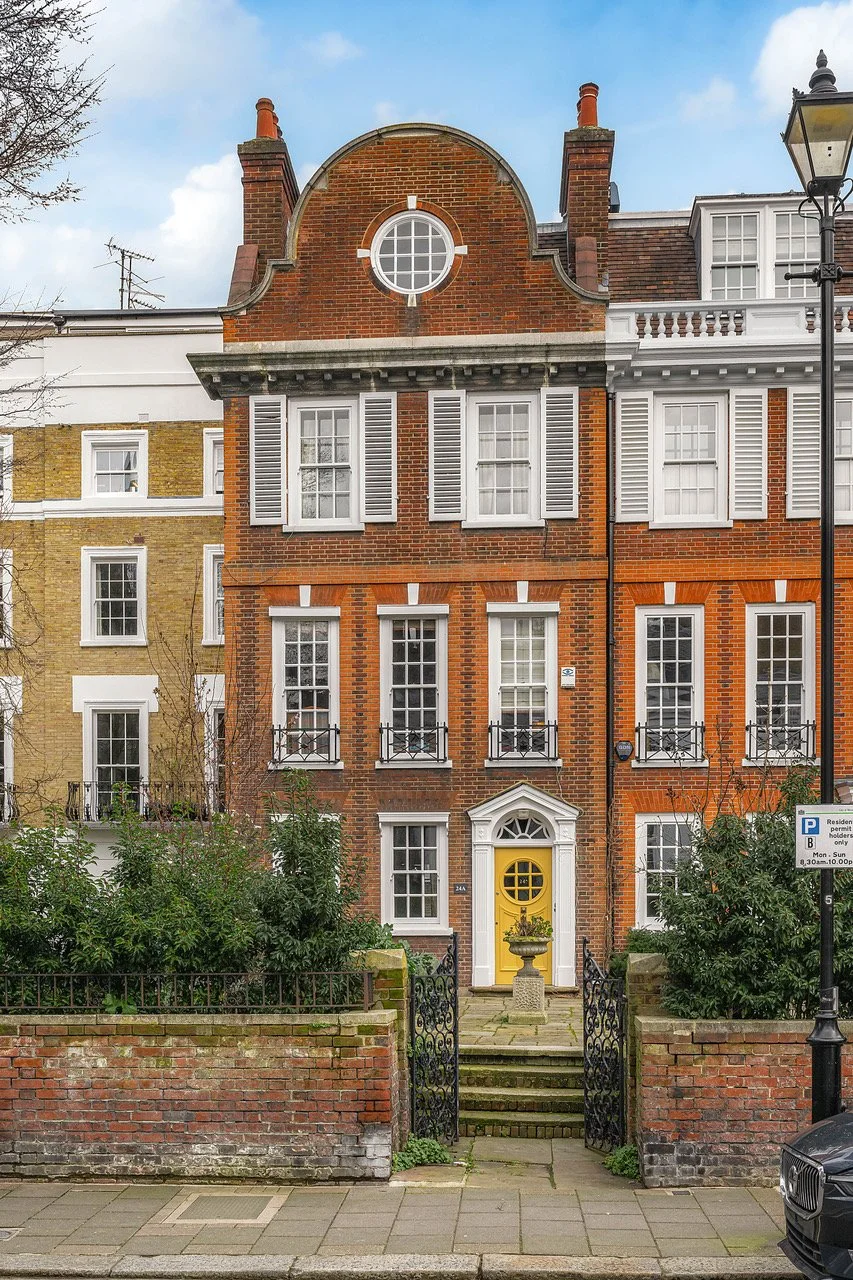 A three-story row house with a yellow front door, white window shutters, and a brick facade. The house has a small front yard with bushes and steps leading to the entrance, enclosed by a short brick wall and black wrought iron gate. A street lamp and parking sign are visible on the sidewalk.