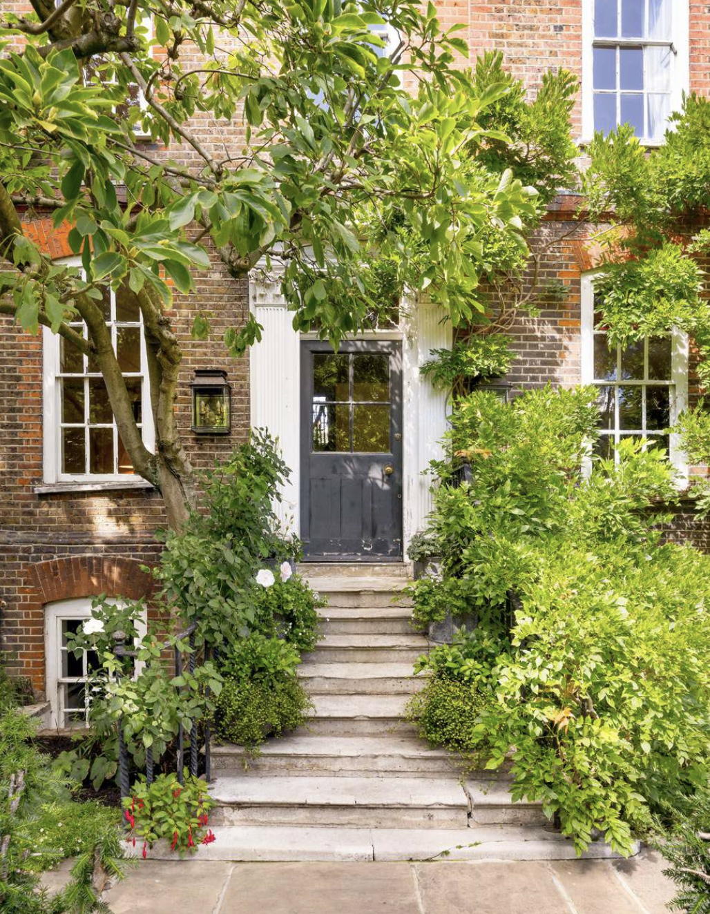 Exterior of a brick house with a black front door, surrounded by lush green plants and trees, with stone steps leading up to the entrance.