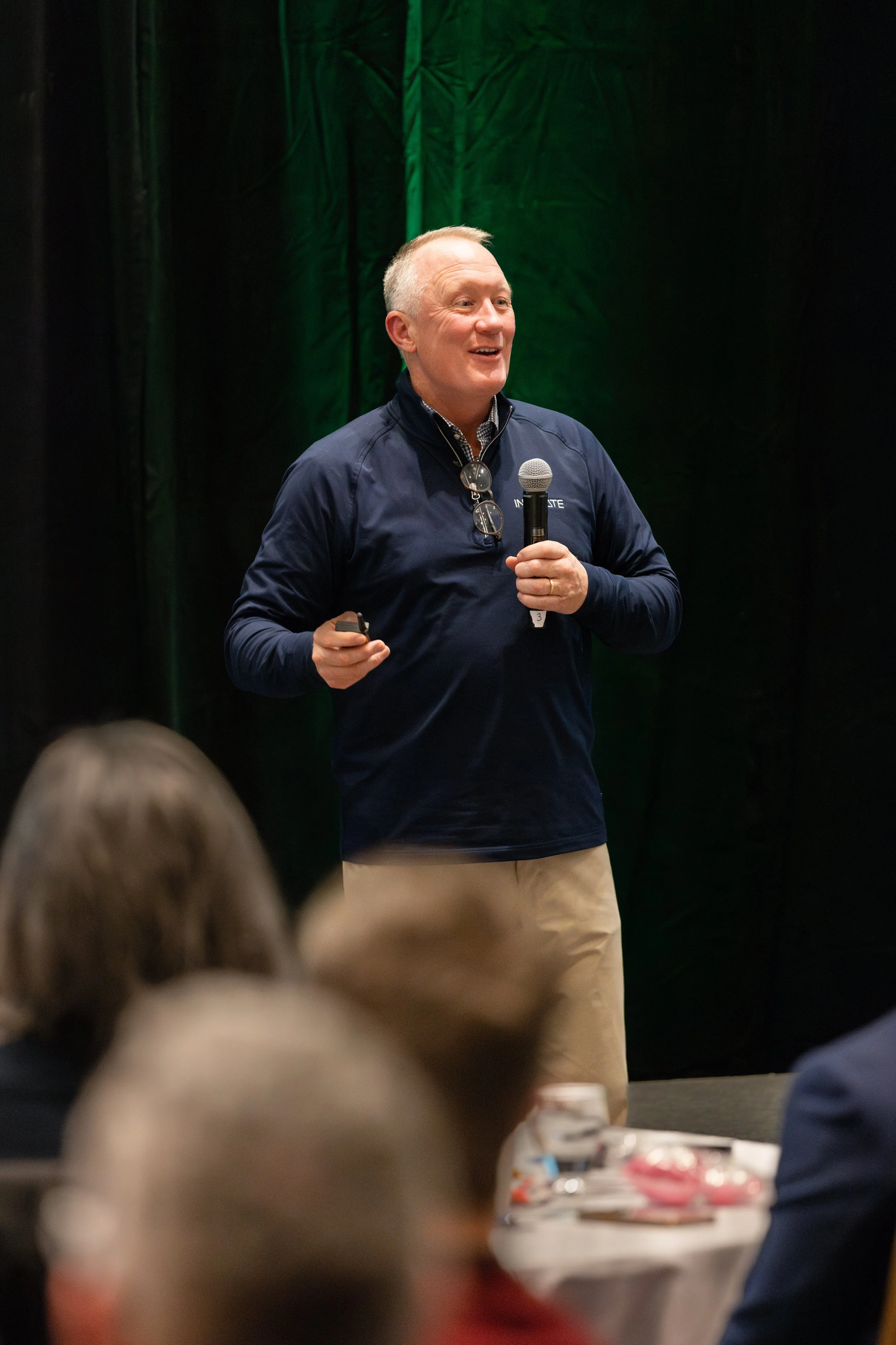 A man giving a presentation at a conference, holding a microphone and a remote, with a dark background and other attendees in the foreground.