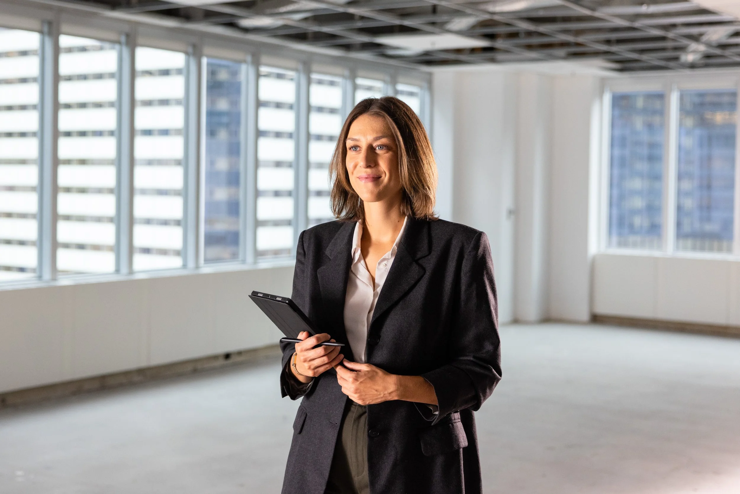A woman in a business suit standing in an empty office space, holding a tablet and looking thoughtfully out the window.
