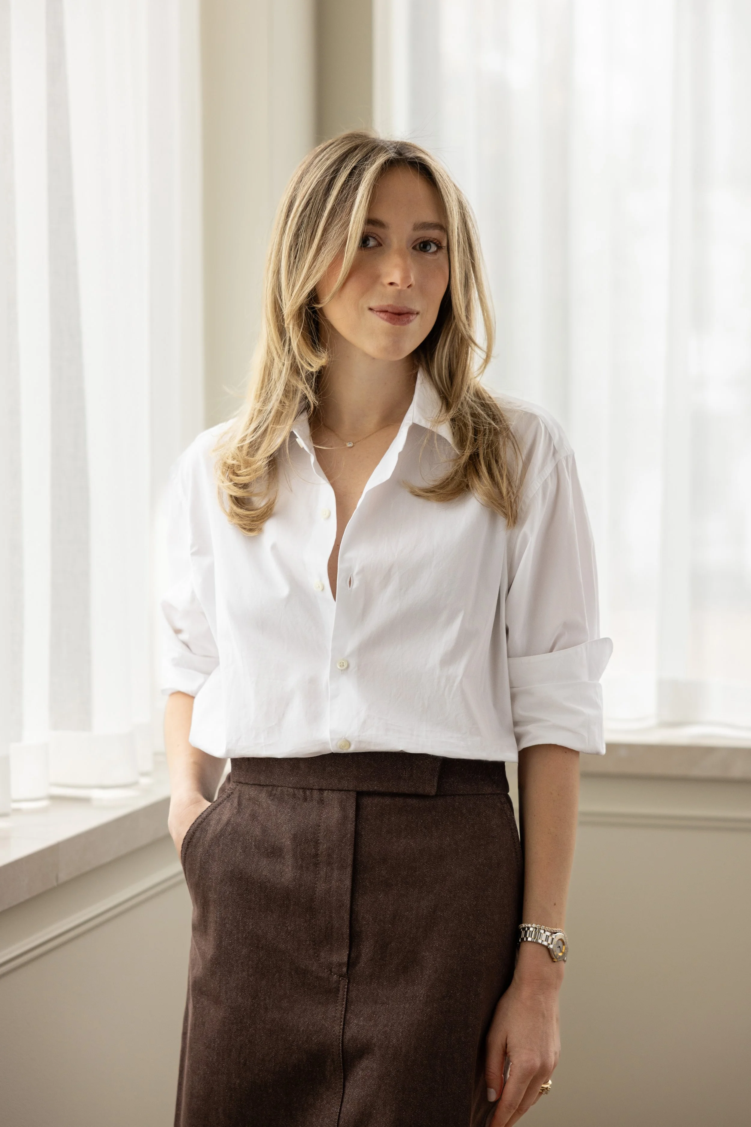 A woman with blonde hair wearing a white button-up shirt and brown skirt standing indoors near a window with white curtains.
