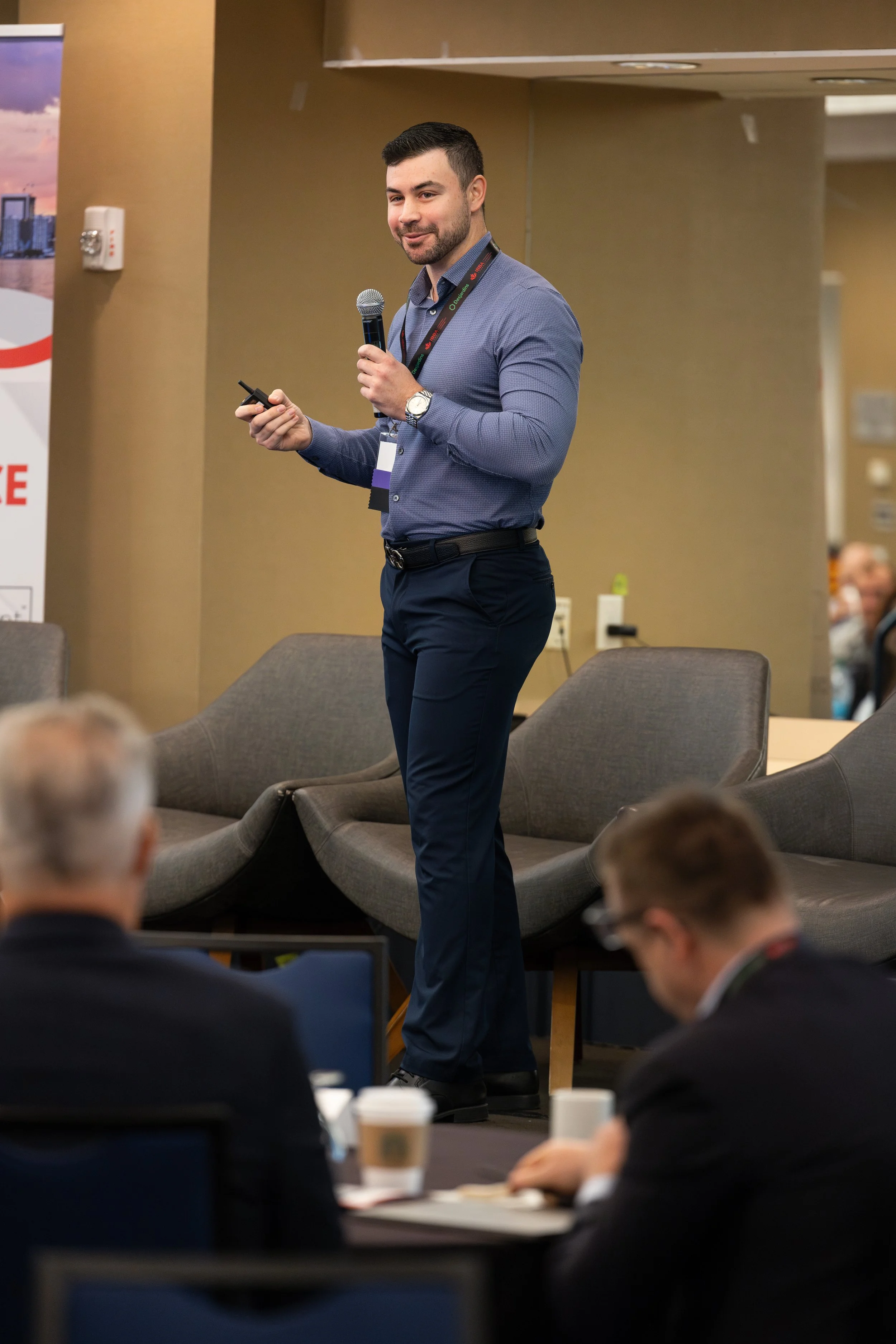 A man in a blue shirt and dark pants delivering a presentation at a conference, holding a microphone and a remote control, with attendees listening and taking notes in the foreground.