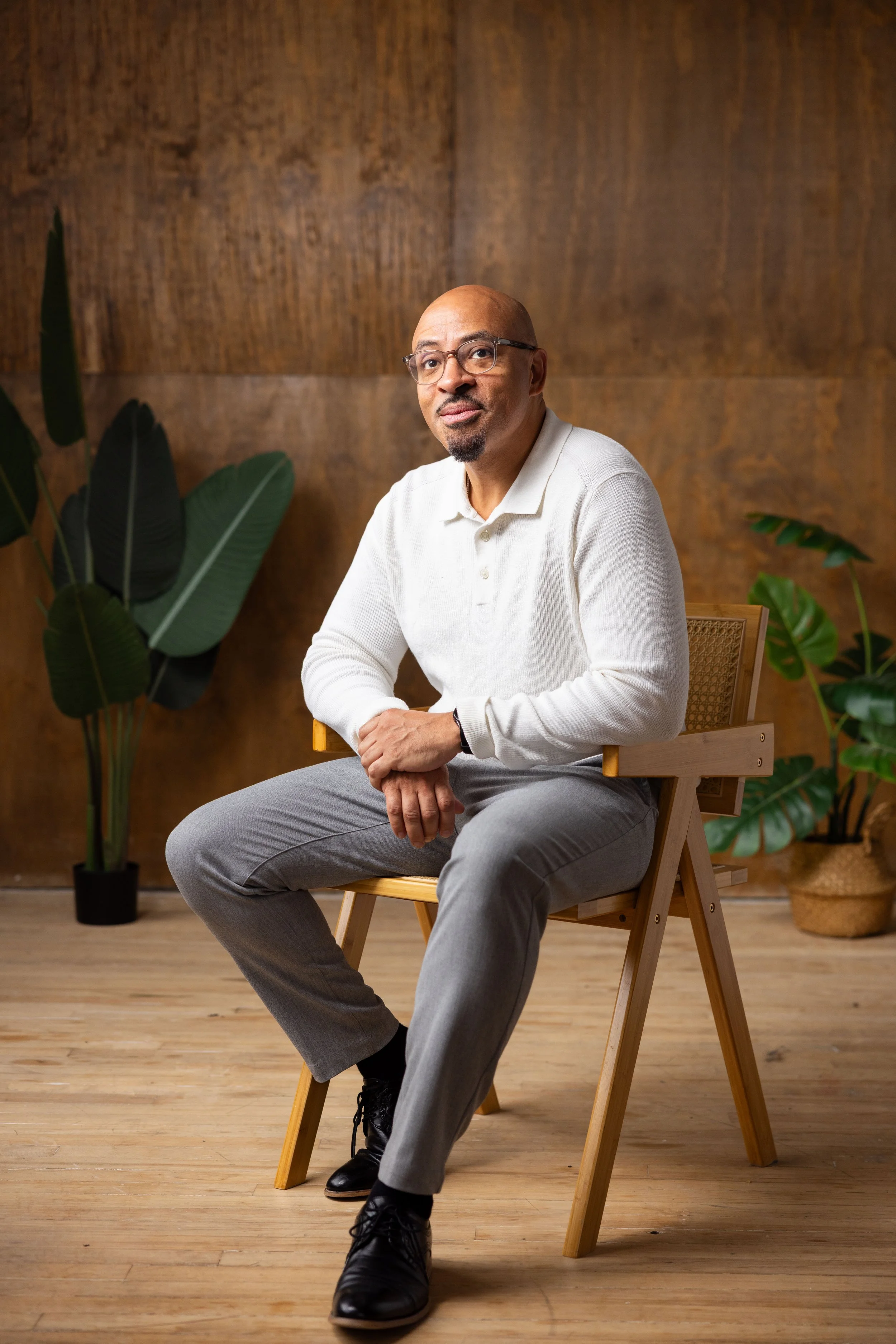 A man sitting on a wooden chair indoors, with a wooden wall and green plants in the background.