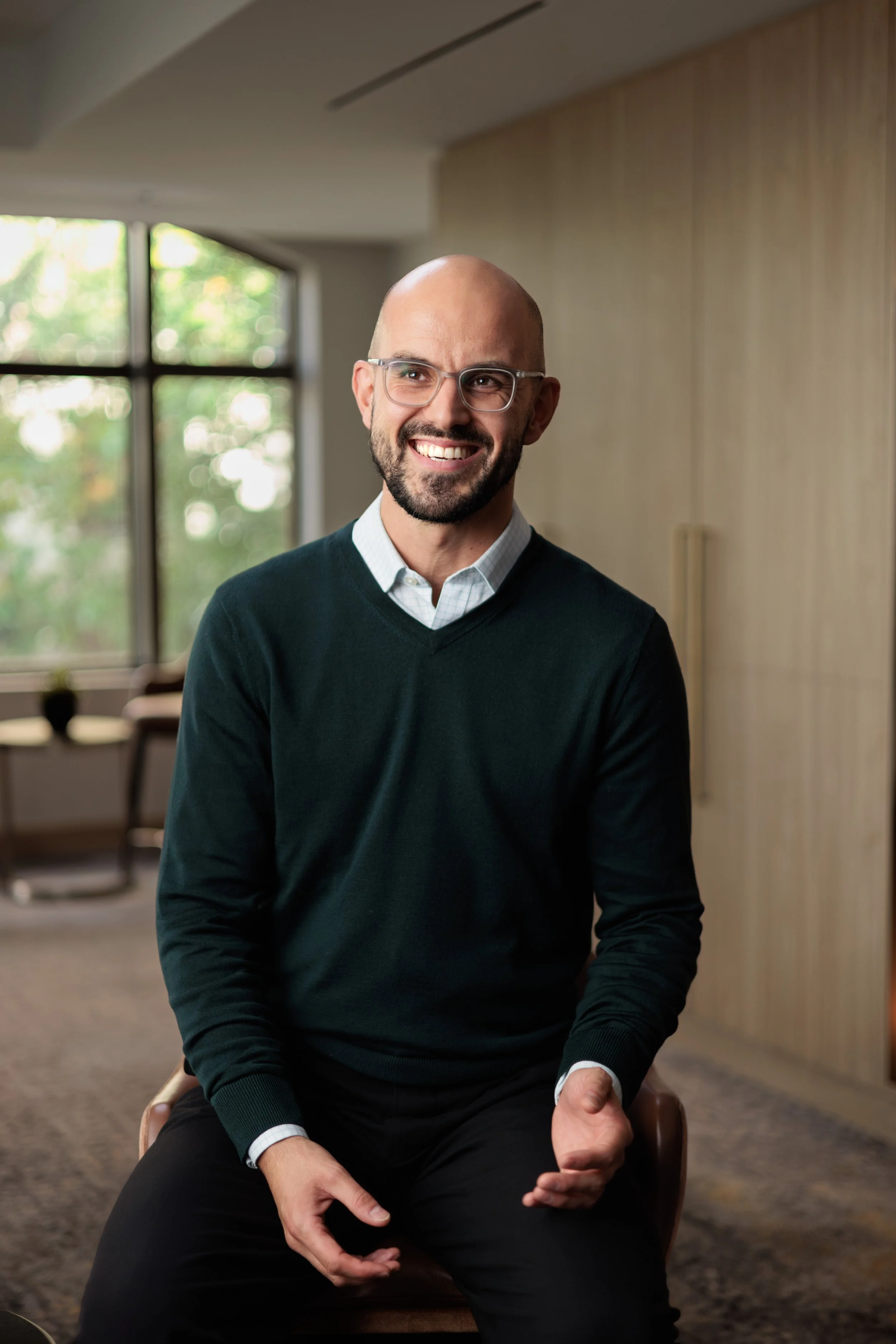 A man with glasses, a beard, and a shaved head, smiling, wearing a dark sweater over a collared shirt, sitting on a chair in a well-lit room with large windows and wooden accents.