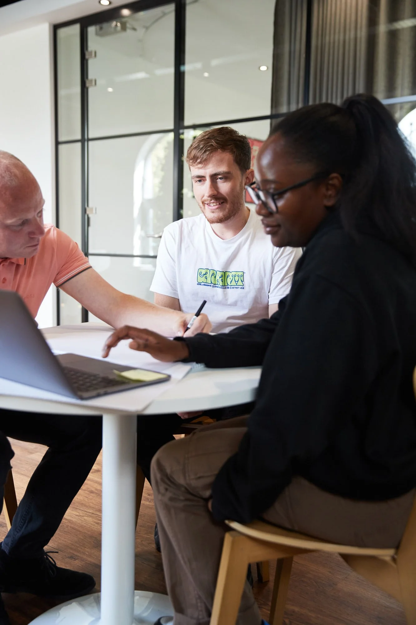 Three people engaged in a discussion around a table with a laptop, papers, and a smartphone.