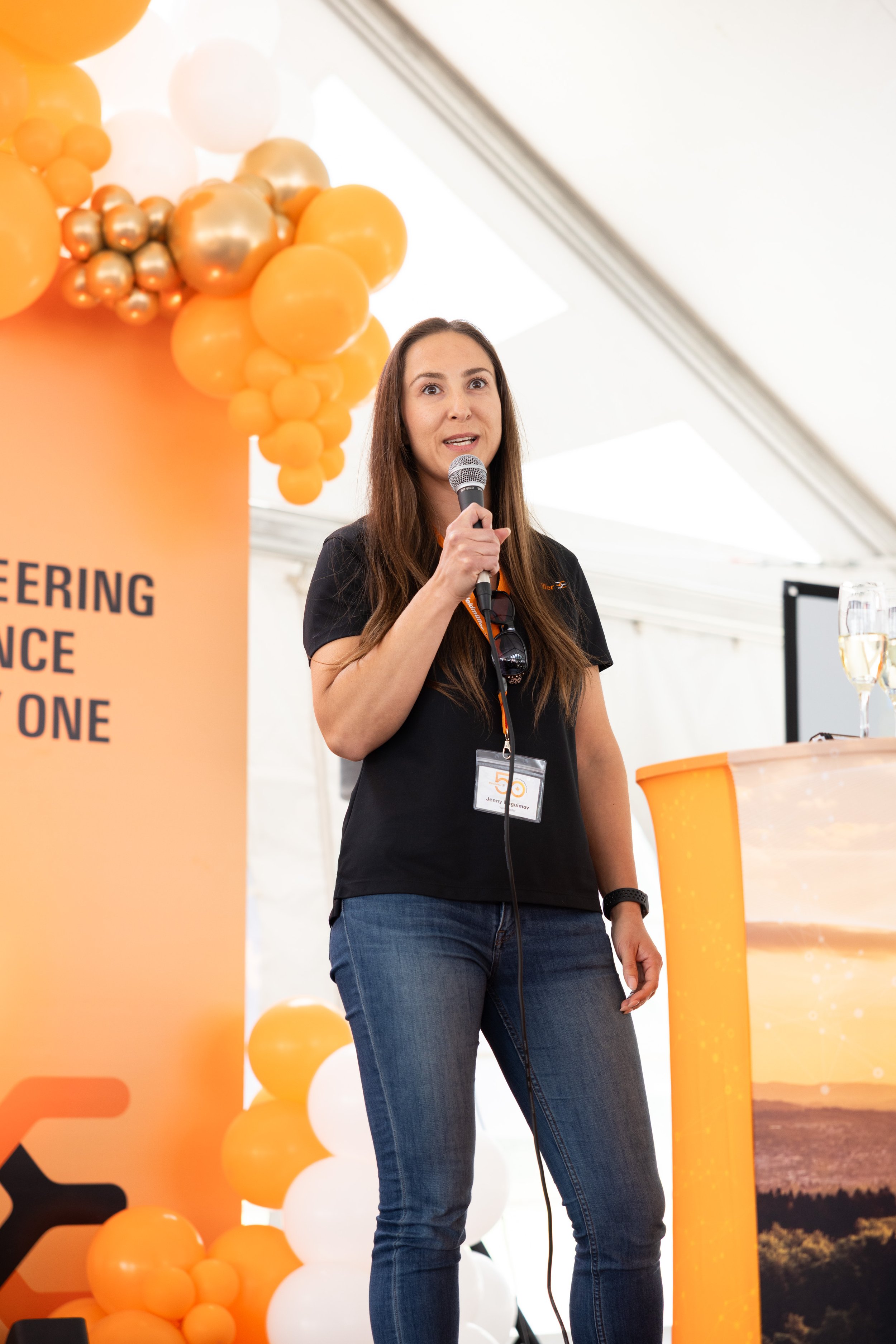 A woman with long brown hair holding a microphone and speaking at a conference or event, standing in front of an orange and white balloon decoration with a sign that reads 'ENGINEERING', 'ADVANCE', and 'ONE'.