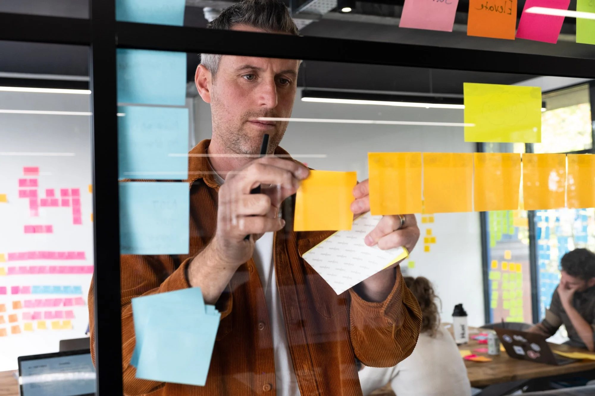 Man in brown jacket writing on yellow sticky notes on glass wall with a notepad in his left hand, in a modern office.