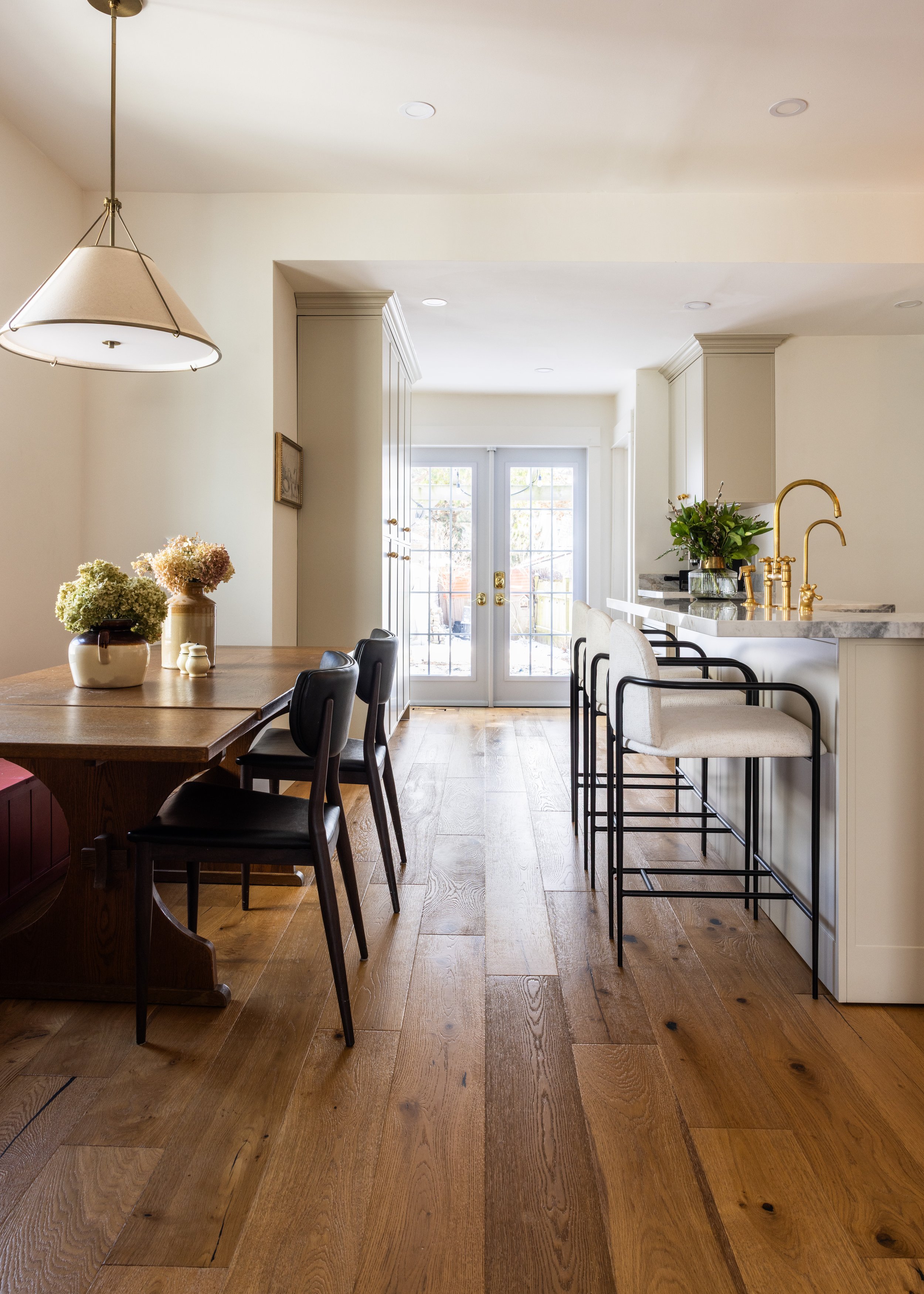 A bright kitchen and dining area with a wooden dining table, black chairs, and a breakfast bar with white stools. Large glass doors lead outside, and the space features hardwood floors, white cabinetry, and gold fixtures.