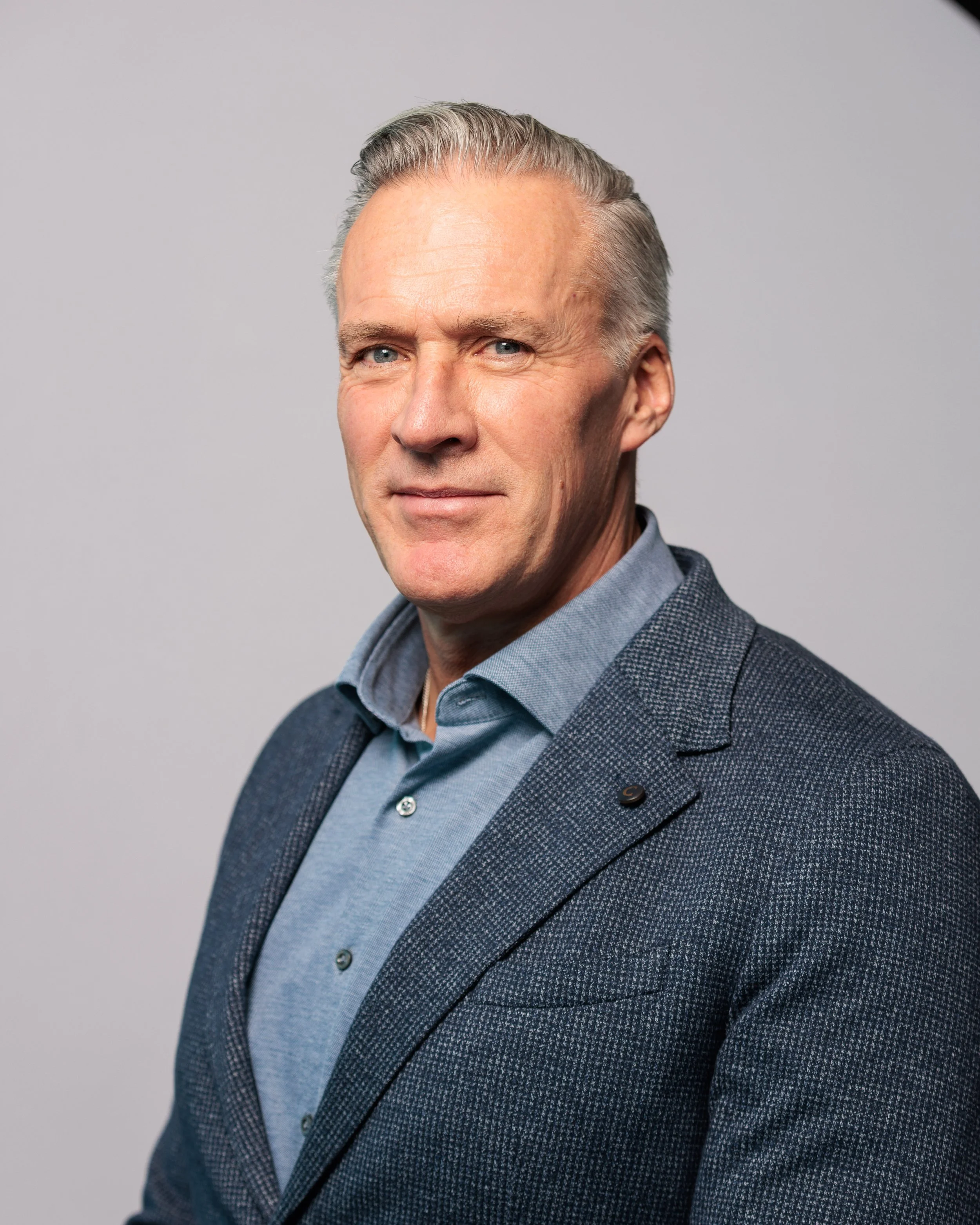 A portrait of a middle-aged man wearing a gray blazer and a blue button-up shirt against a plain gray background.