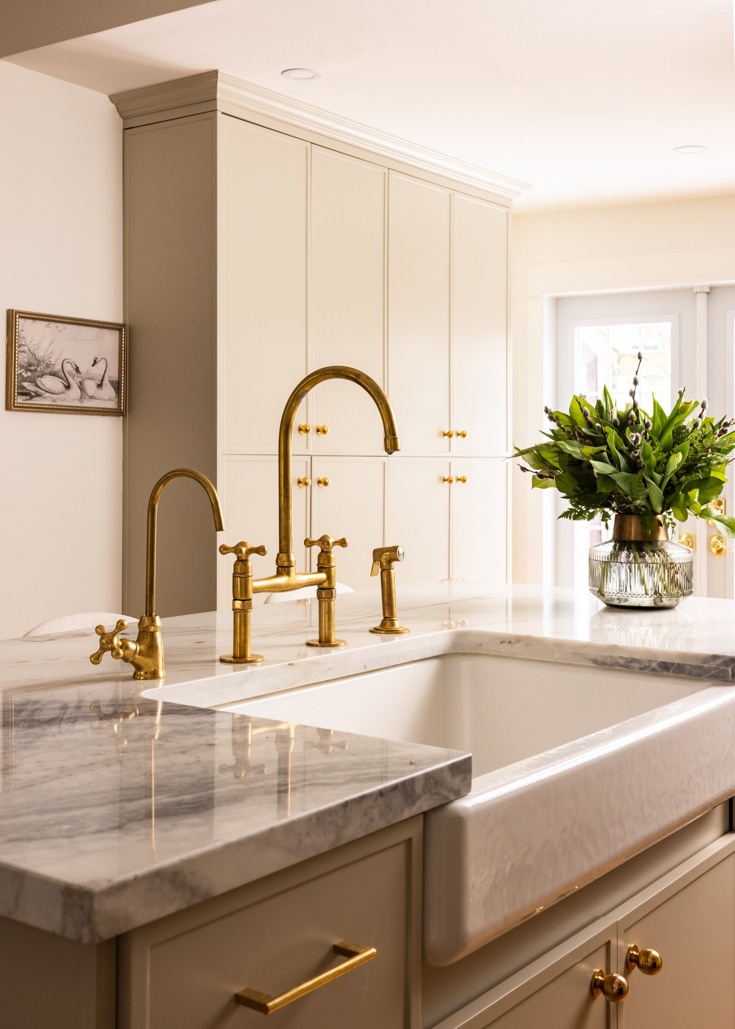 A kitchen sink with a marble countertop, brass faucets, and a large vase with a green plant, with white cabinets and a window in the background.