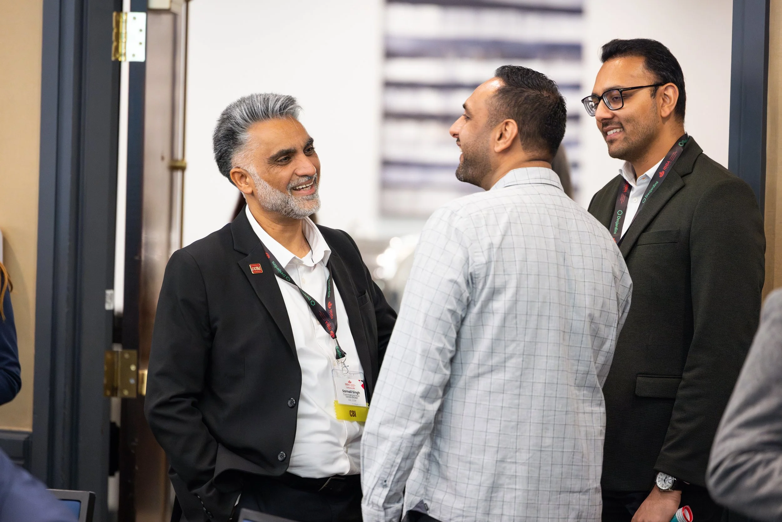 Three men in business attire engaging in conversation at a professional event. One man has gray hair and a beard, wearing a black suit and white shirt. The second man has short black hair and is wearing a checked shirt. The third man has black hair, 