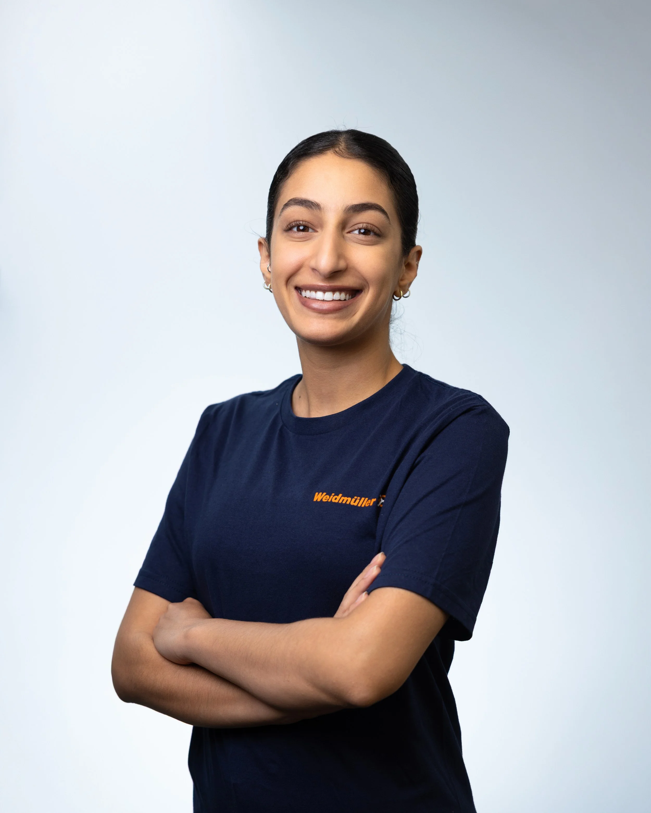 A young woman with dark hair tied back, smiling, wearing a navy blue t-shirt with orange text on the chest, arms crossed, standing against a plain light grey background.