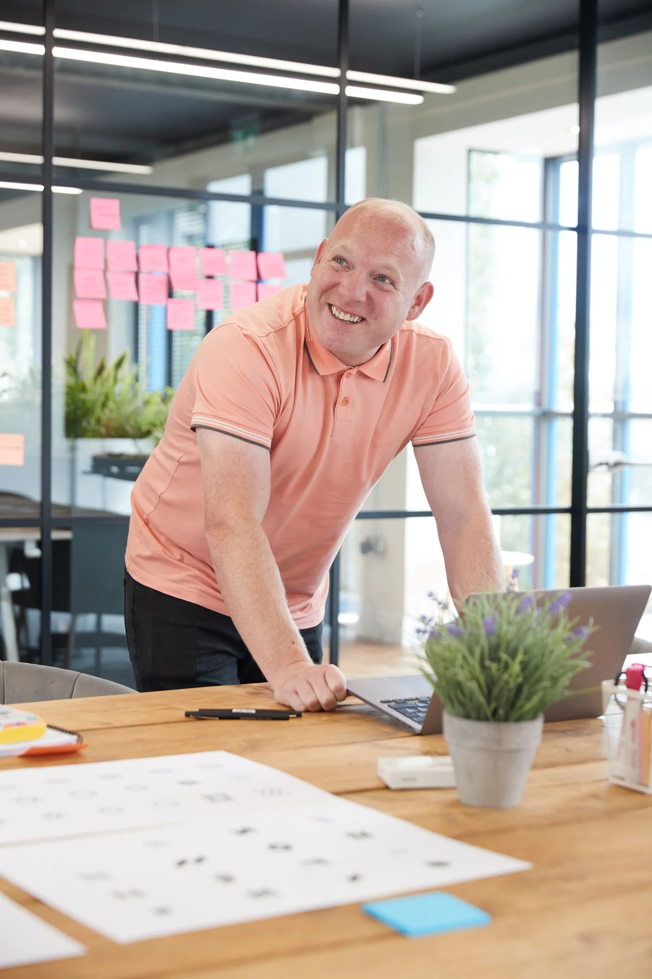 A man in a peach polo shirt smiling and leaning on a laptop on a wooden table in a bright office with large windows and pink sticky notes on the glass wall behind him.