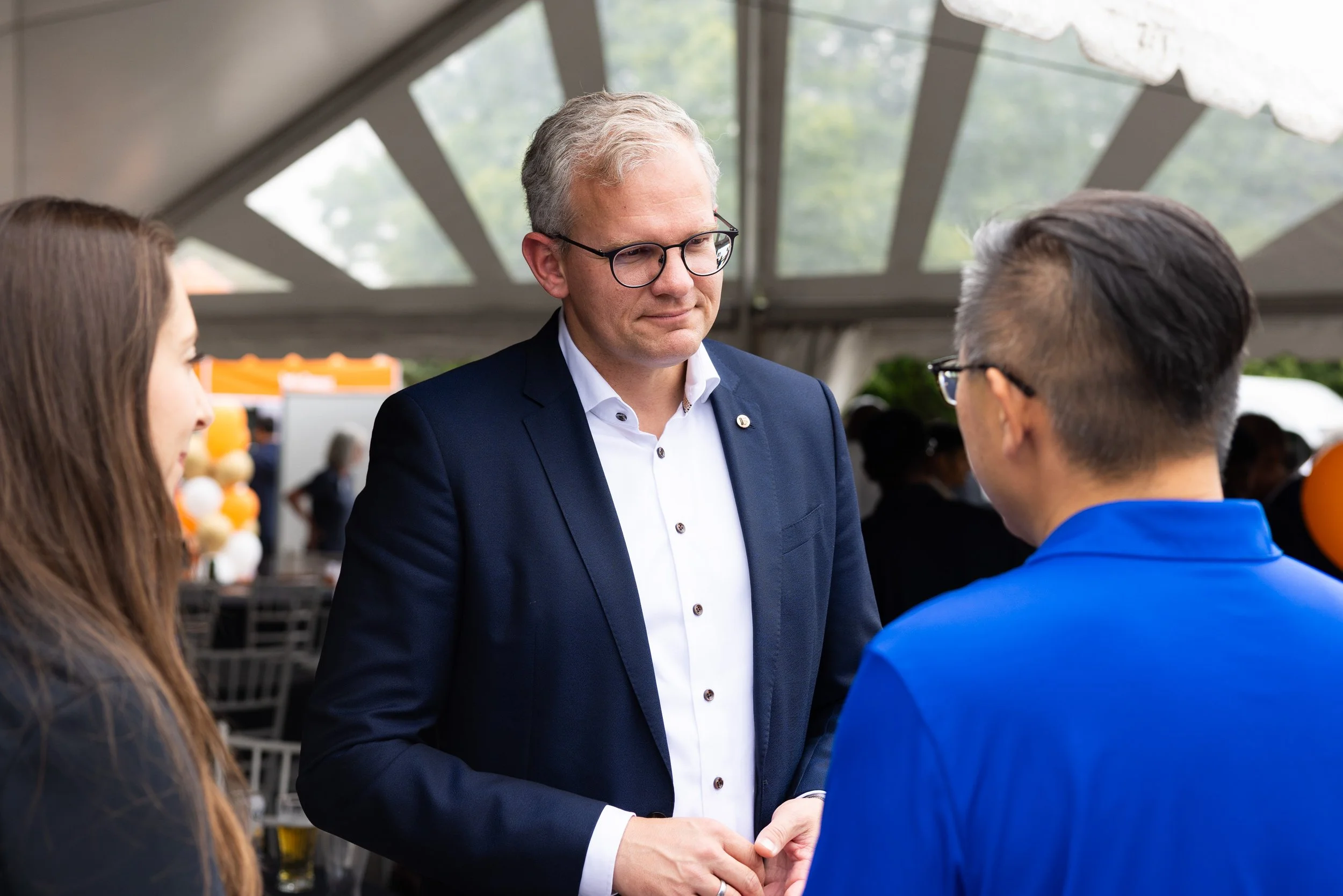 A man in a navy suit and glasses talking to a woman and a man in a blue jacket inside a tent at an event.