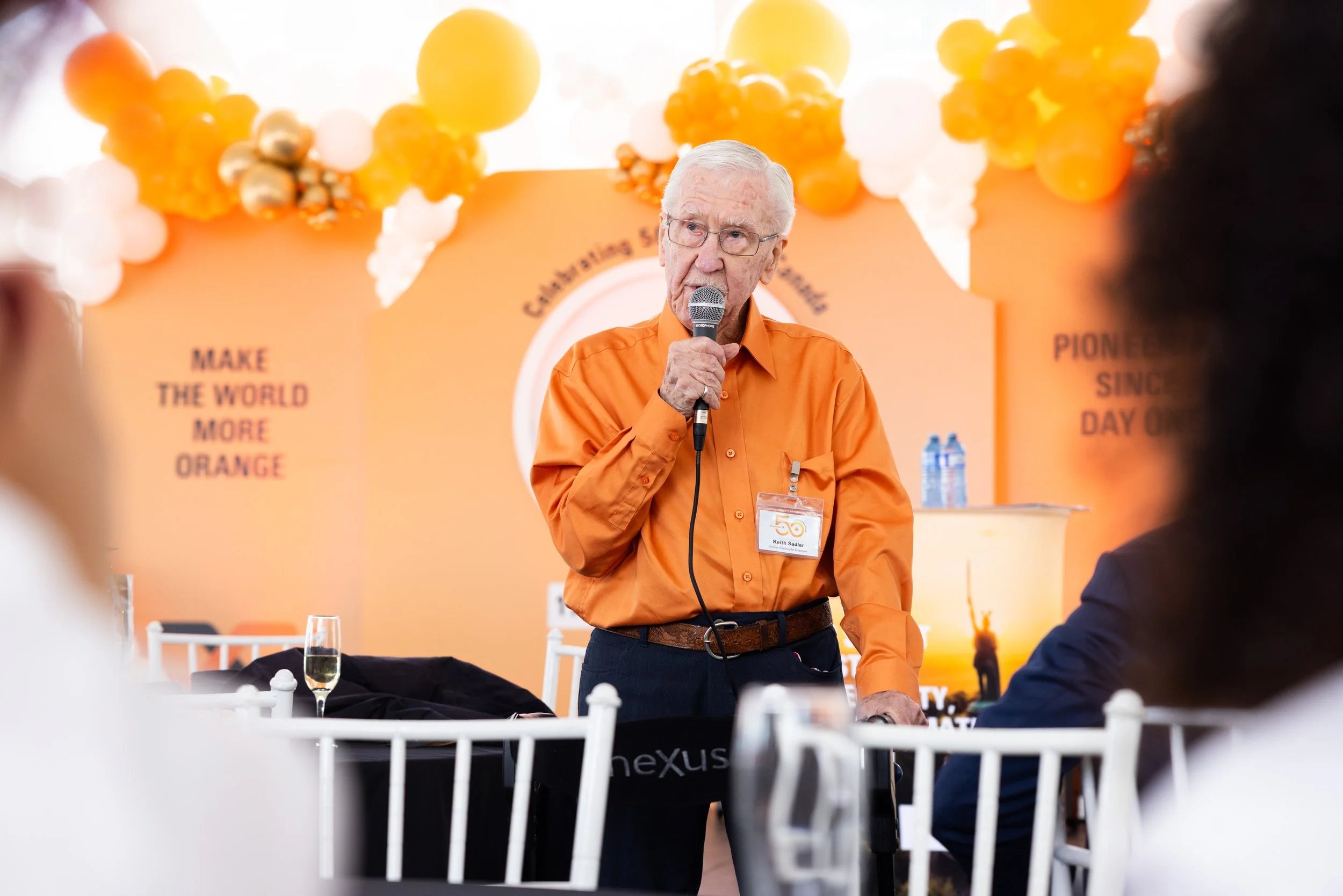 An elderly man with white hair and glasses, dressed in an orange shirt, holding a microphone, speaking at a conference or event with a peach-colored background decorated with orange and white balloons. There are people seated in front of him.