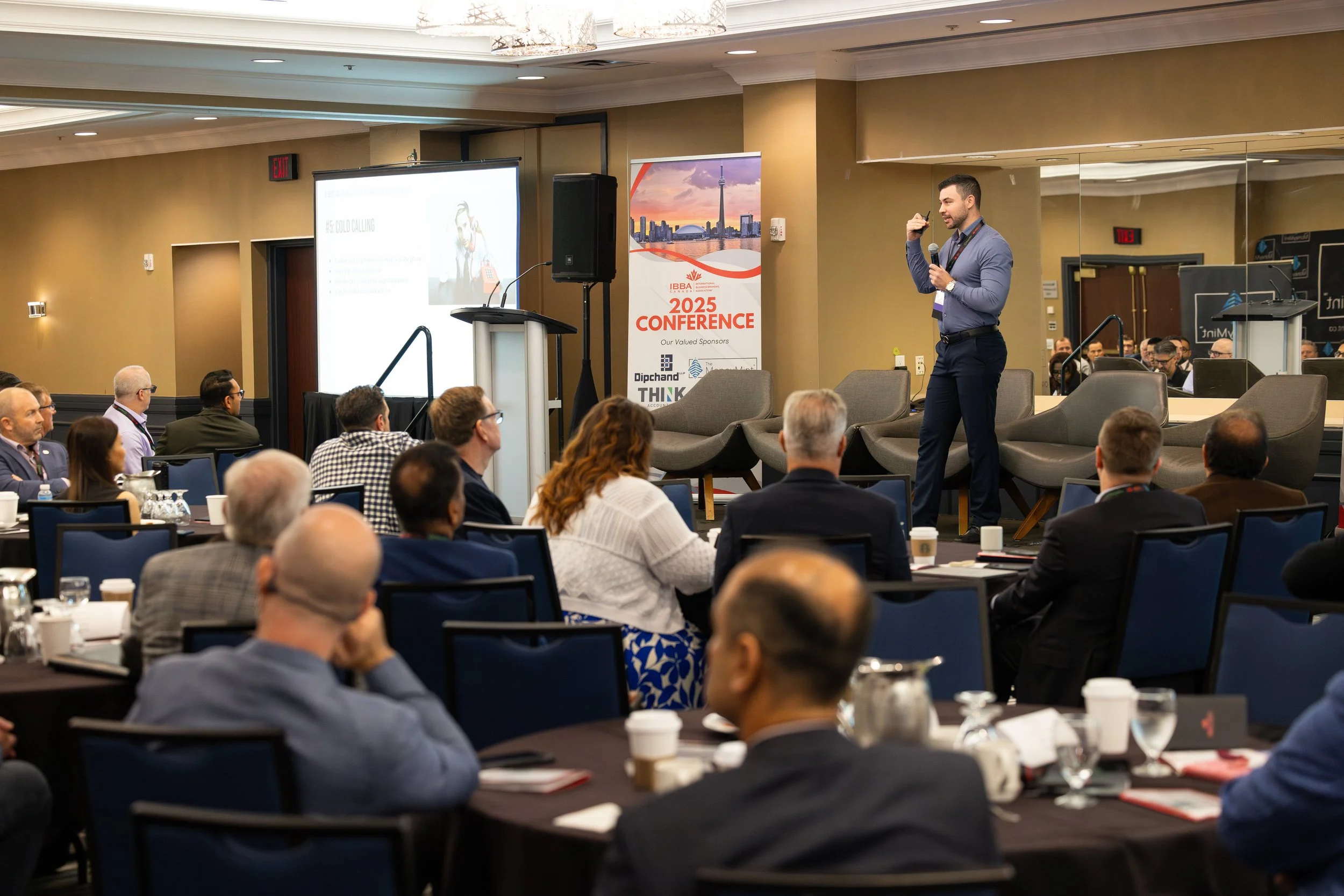 A speaker giving a presentation at a conference, with an audience seated at round tables. There is a large screen displaying slides and a banner that reads 'IBBA 2025 Conference' in the background.
