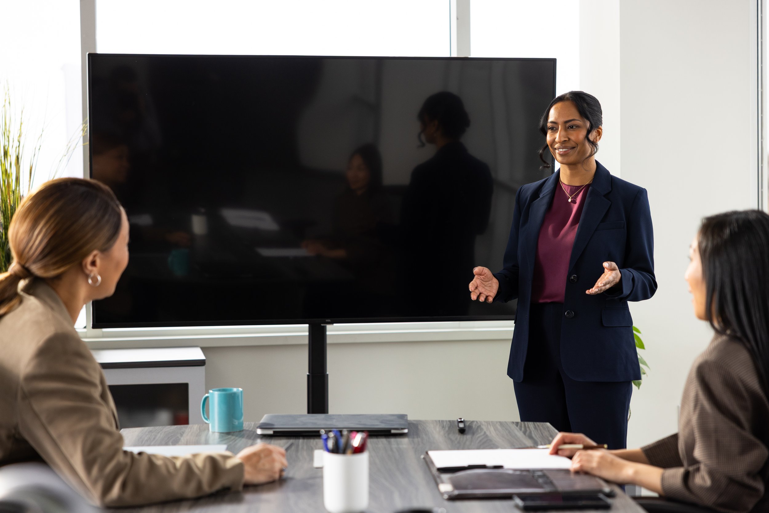 A woman in a navy blazer giving a presentation to three women seated at a conference table in a modern office, with a large black screen behind her.