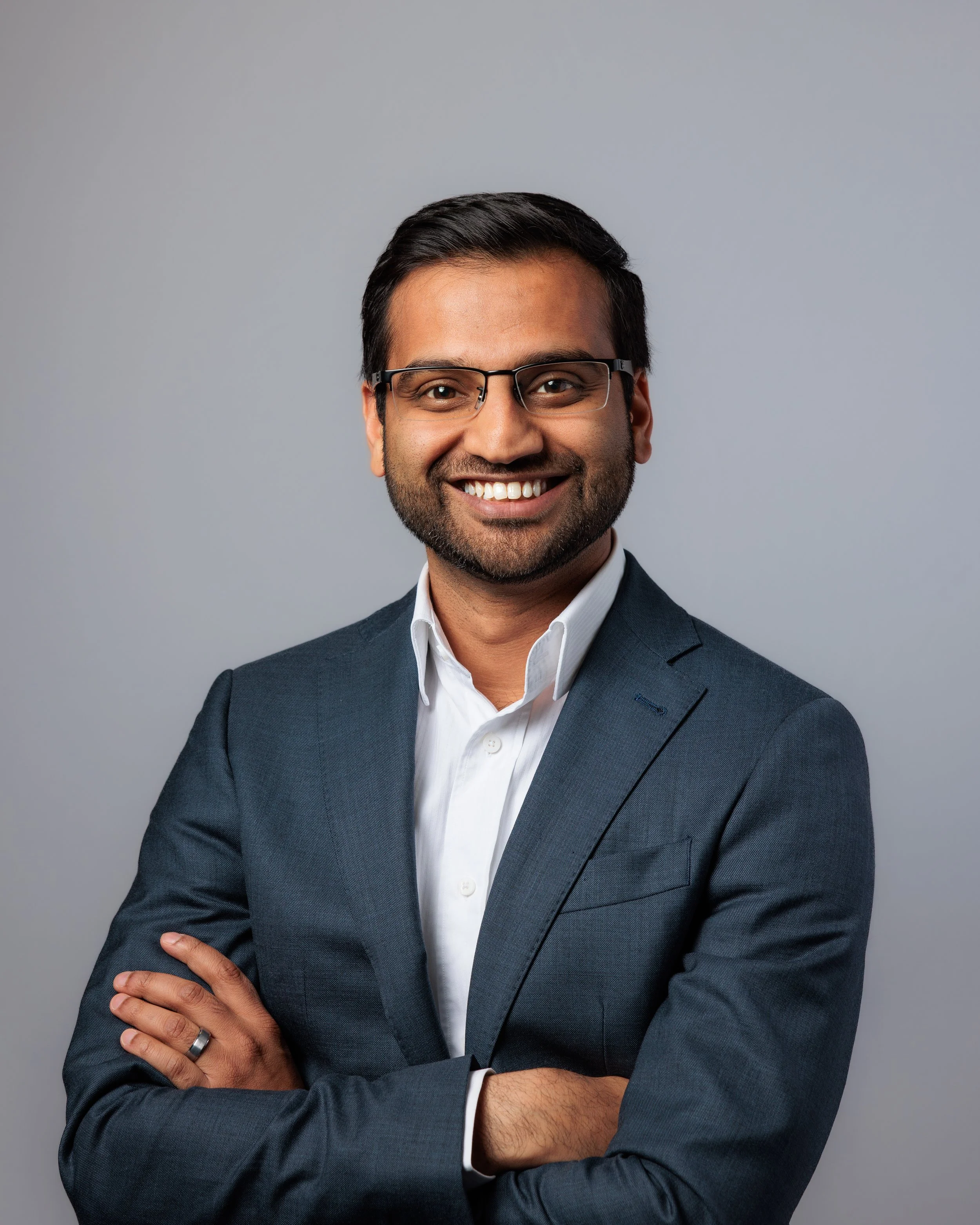 A man in a dark gray business suit and white shirt, smiling with arms crossed, standing against a light gray background.