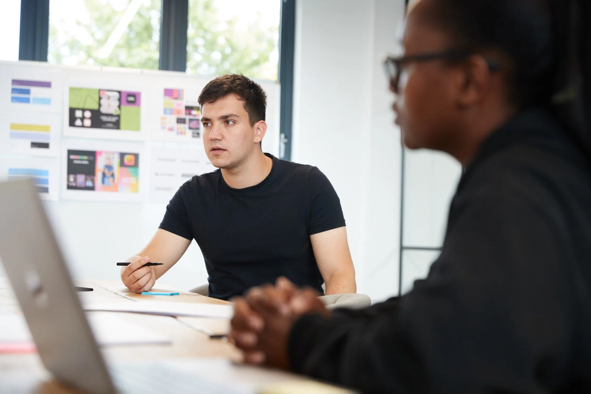 Two men in a professional meeting or discussion, one young man in a black T-shirt holding a pen, the other man in a dark jacket, with a blurred background of charts and designs on a whiteboard or wall.