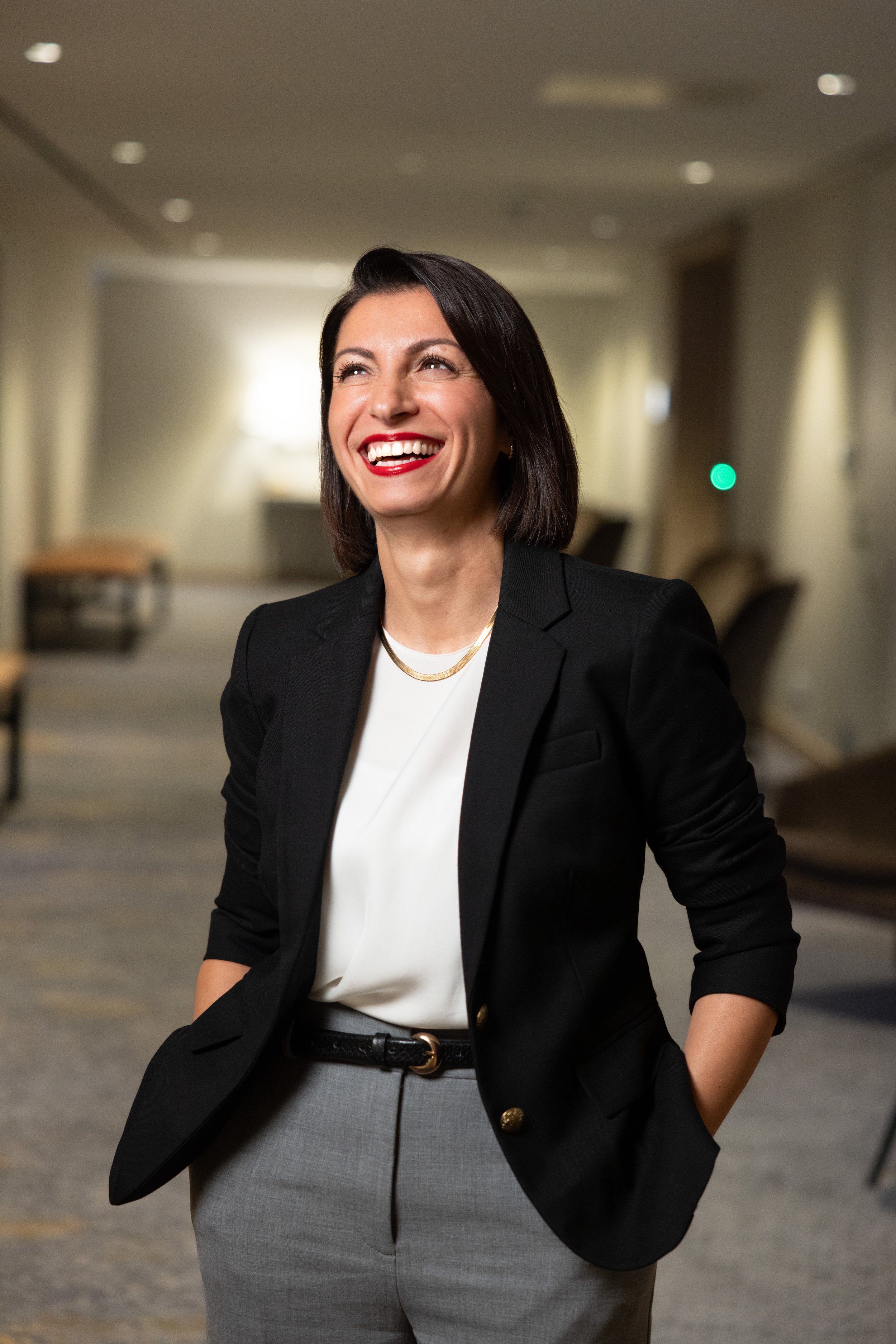 A woman with dark hair smiling, dressed in a black blazer, white top, and grey pants, standing in a well-lit indoor setting.