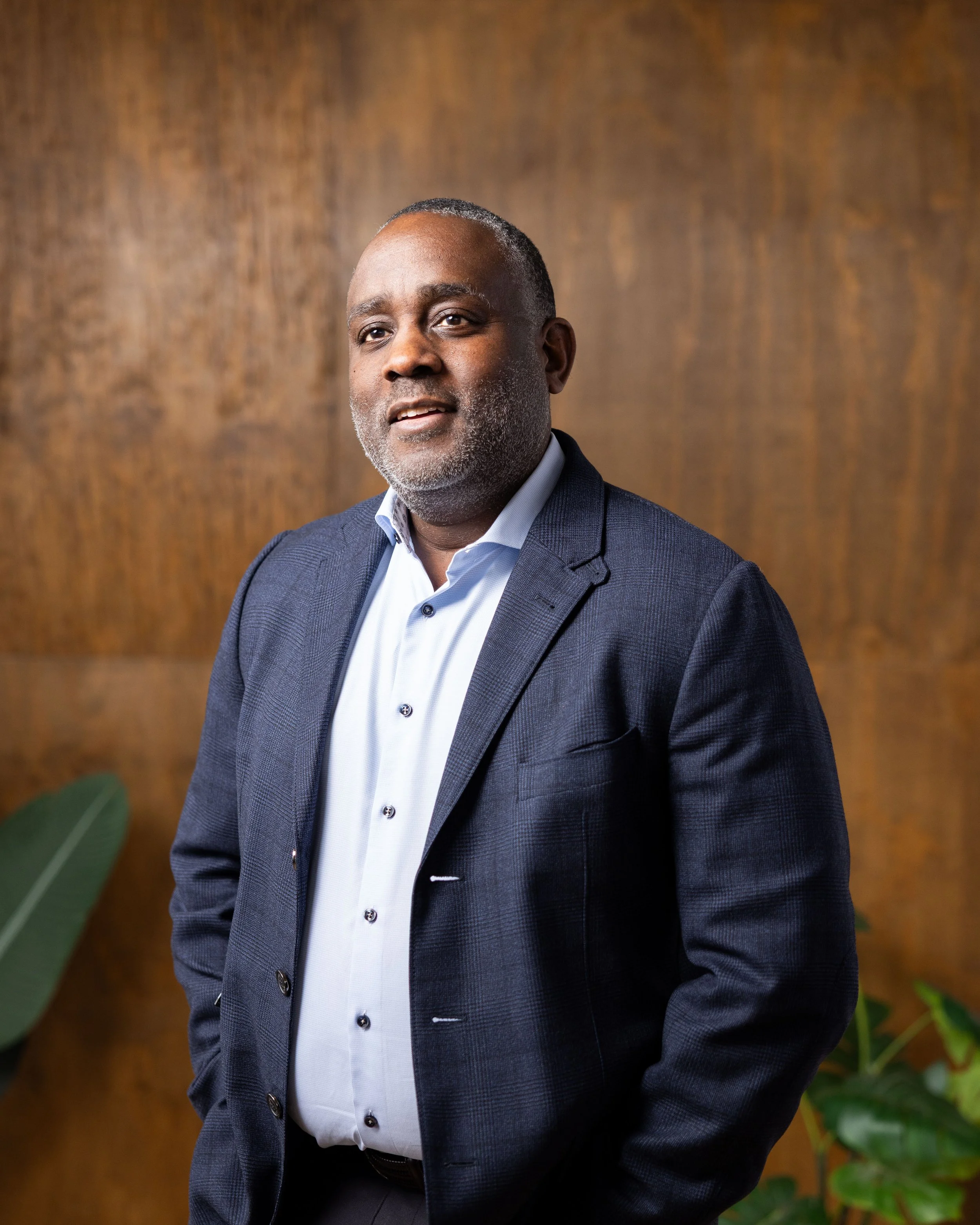 A middle-aged Black man wearing a navy blazer and light blue shirt, standing indoors with a wood-paneled wall and green plants in the background.