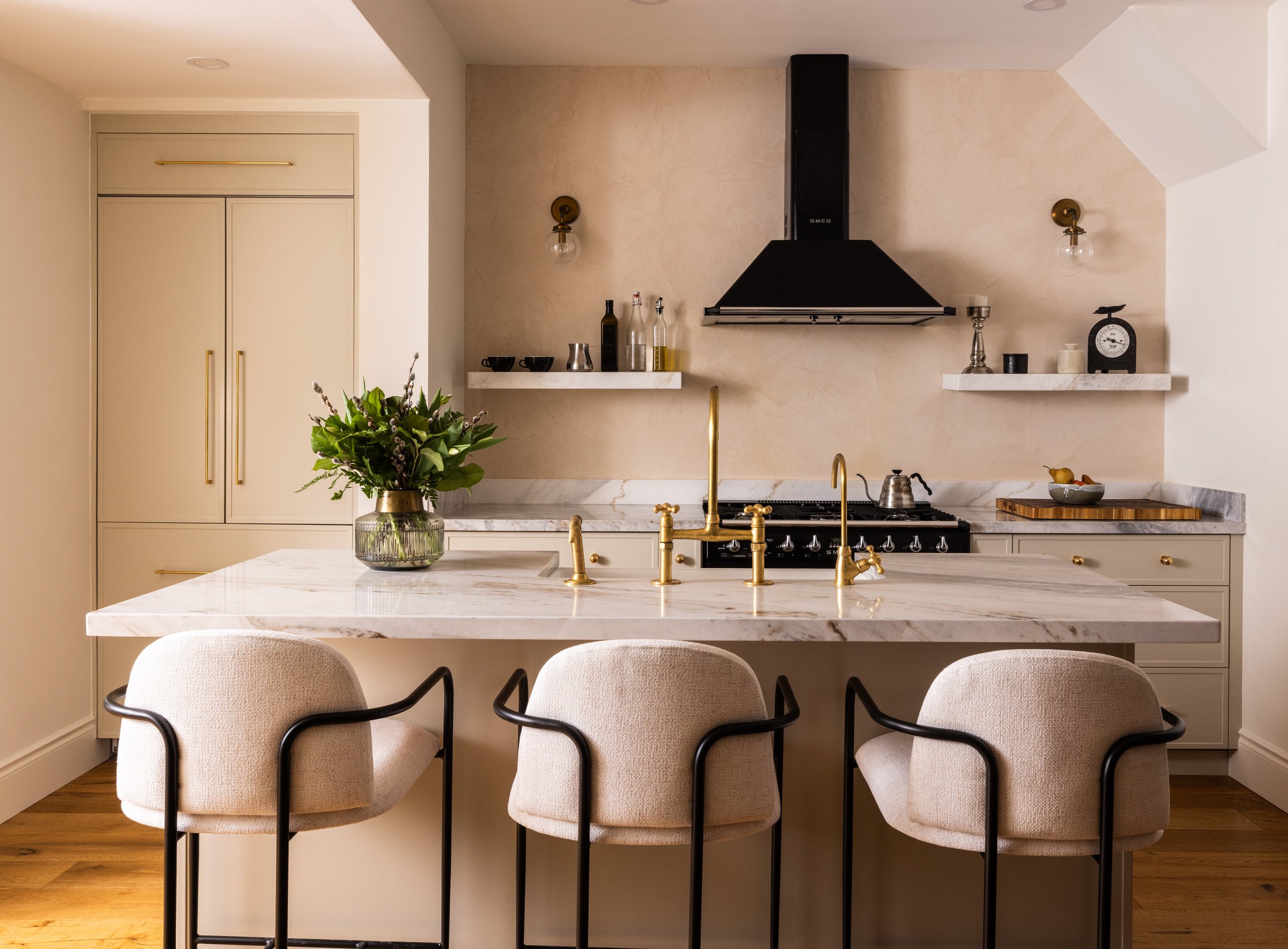 Modern kitchen with a marble island, three beige upholstered chairs with black frames, and gold accents. Features a black range hood, open white shelves with kitchen decor, and a bouquet of flowers in a glass vase on the island. Wooden flooring and n
