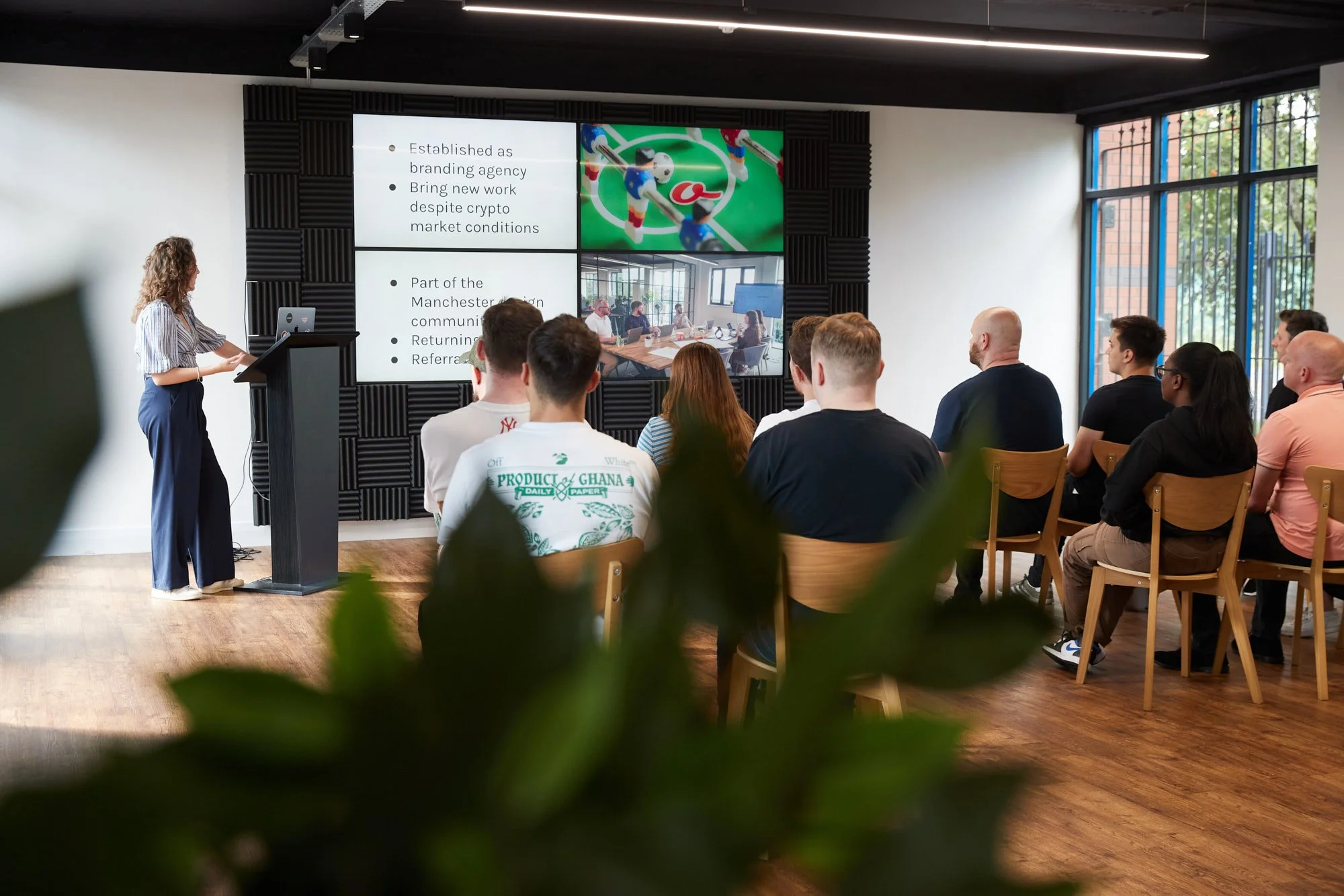 A woman giving a presentation to an audience in a modern conference room with large windows, a big screen displaying a slide with text and images, and a wooden floor. The audience is seated facing the presenter and screen.