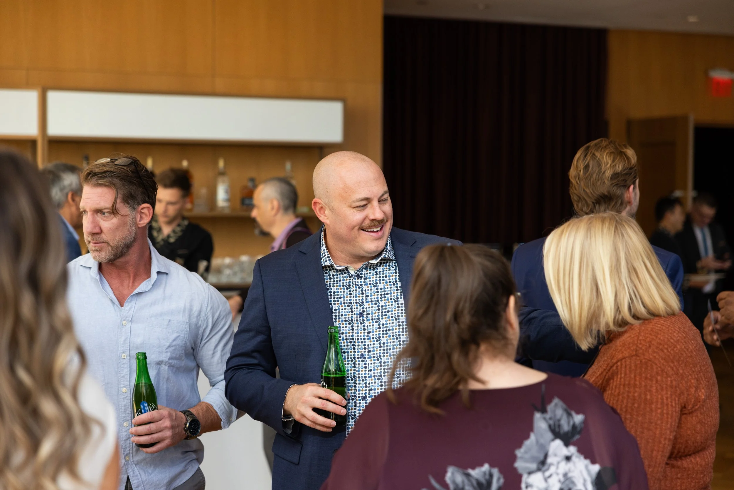 A group of people socializing at a bar or party, with two men in the foreground holding drinks and talking.