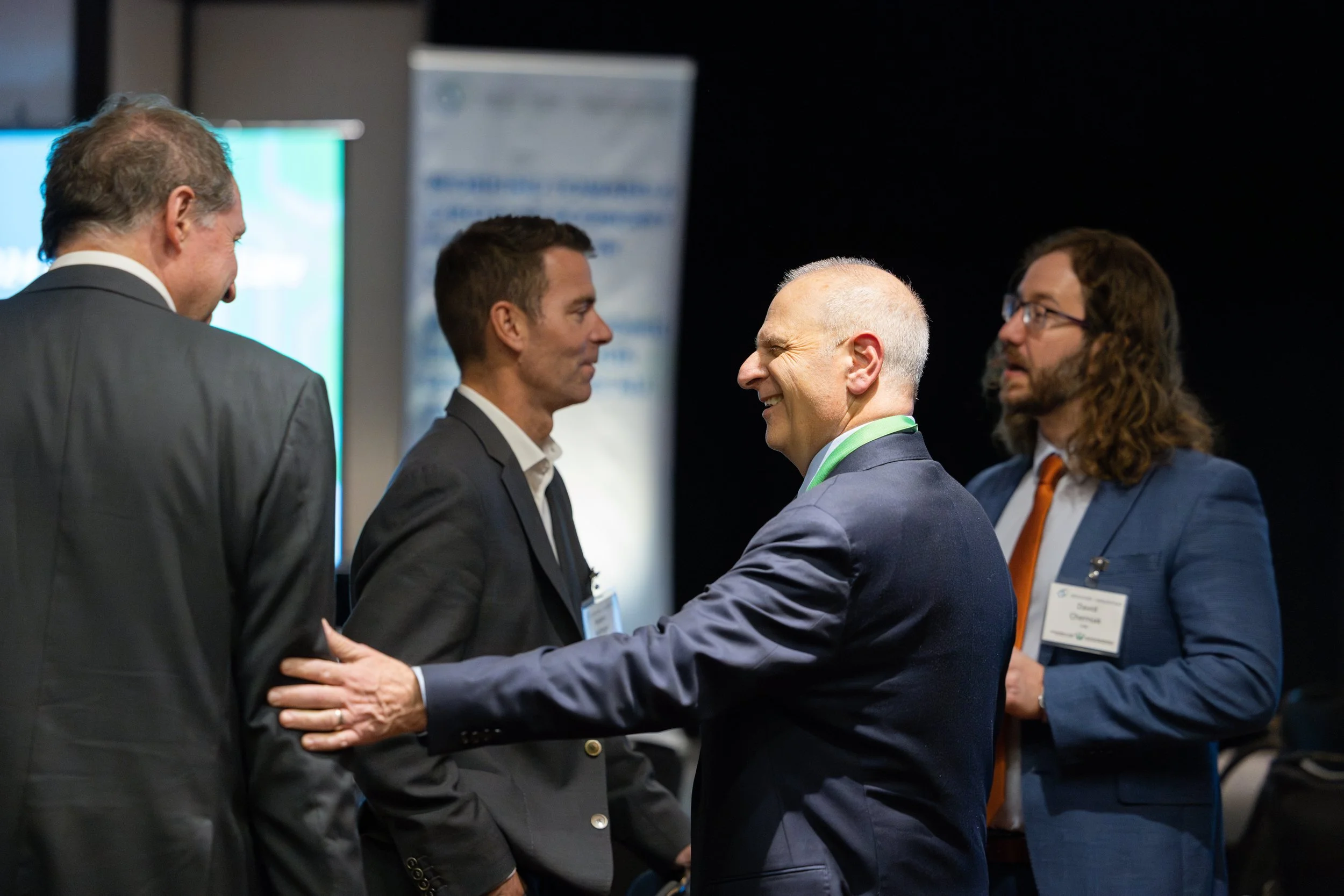 Four men in suits engaged in conversation at a professional event or conference, with badges and a presentation screen in the background.