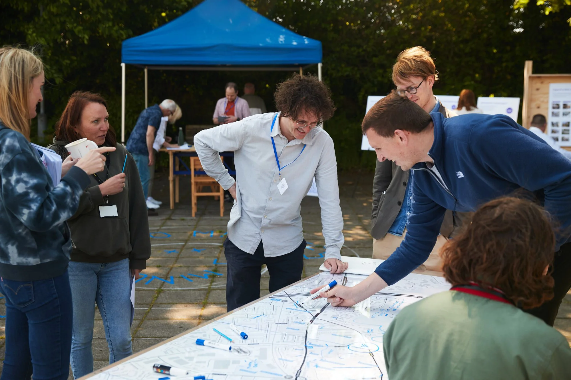 Group of people gathered around a table outdoors, discussing plans marked on a large paper map during a daytime event.