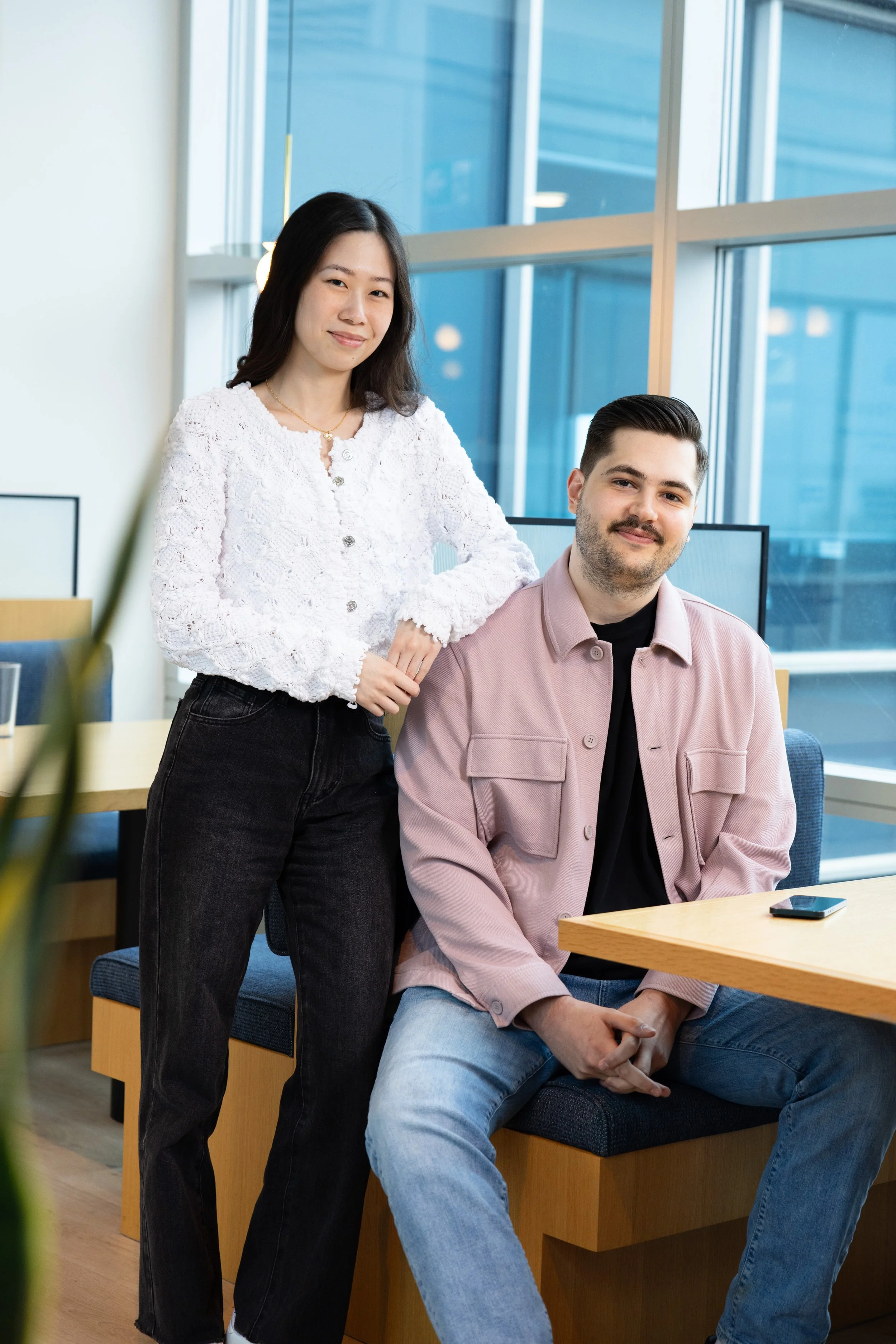Two young adults, a woman and a man, in a modern office space. The woman is standing, dressed in a white textured top and black pants, with her hand resting on the seated man's shoulder. The man is sitting, wearing a pink jacket over a black shirt, w
