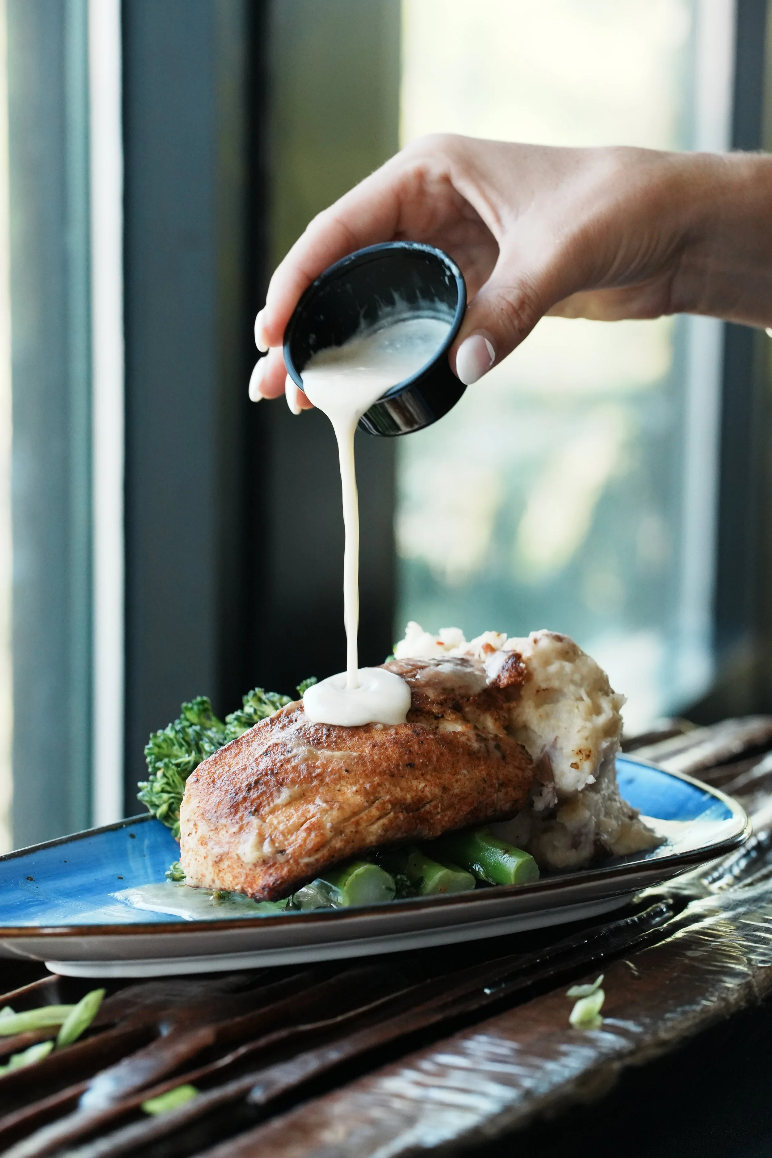 A person pouring white gravy over a serving of chicken with mashed potatoes and green beans on a blue plate.
