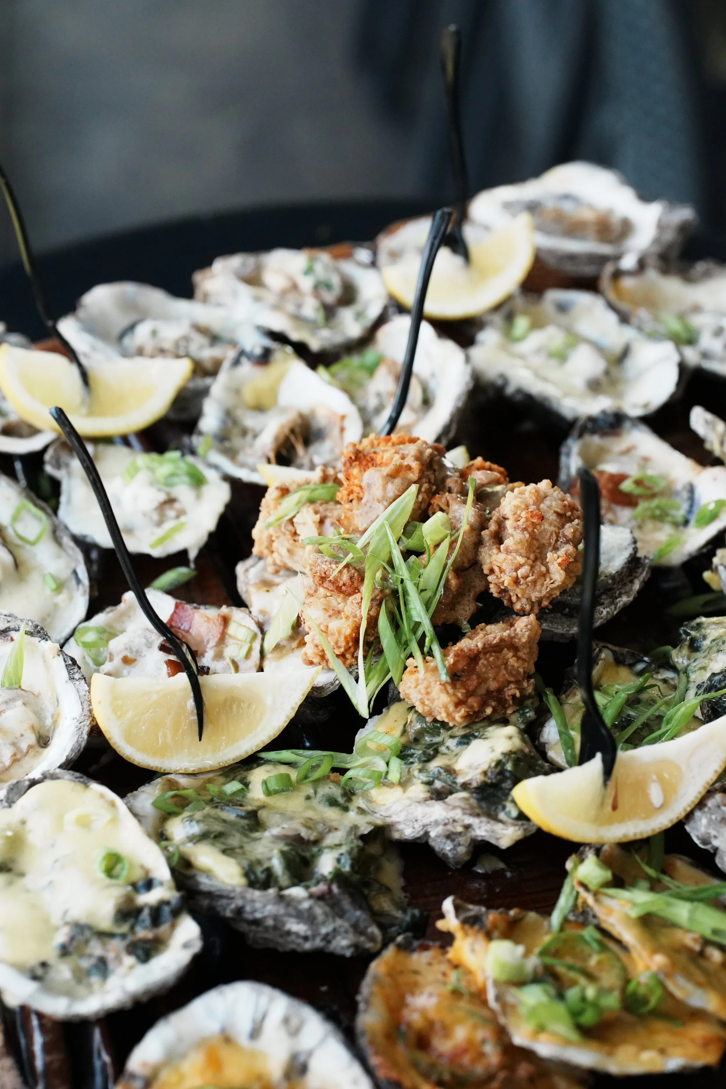 A platter of fresh oysters garnished with lemon wedges, chopped green onions, and a fried oyster or chicken strip on top.