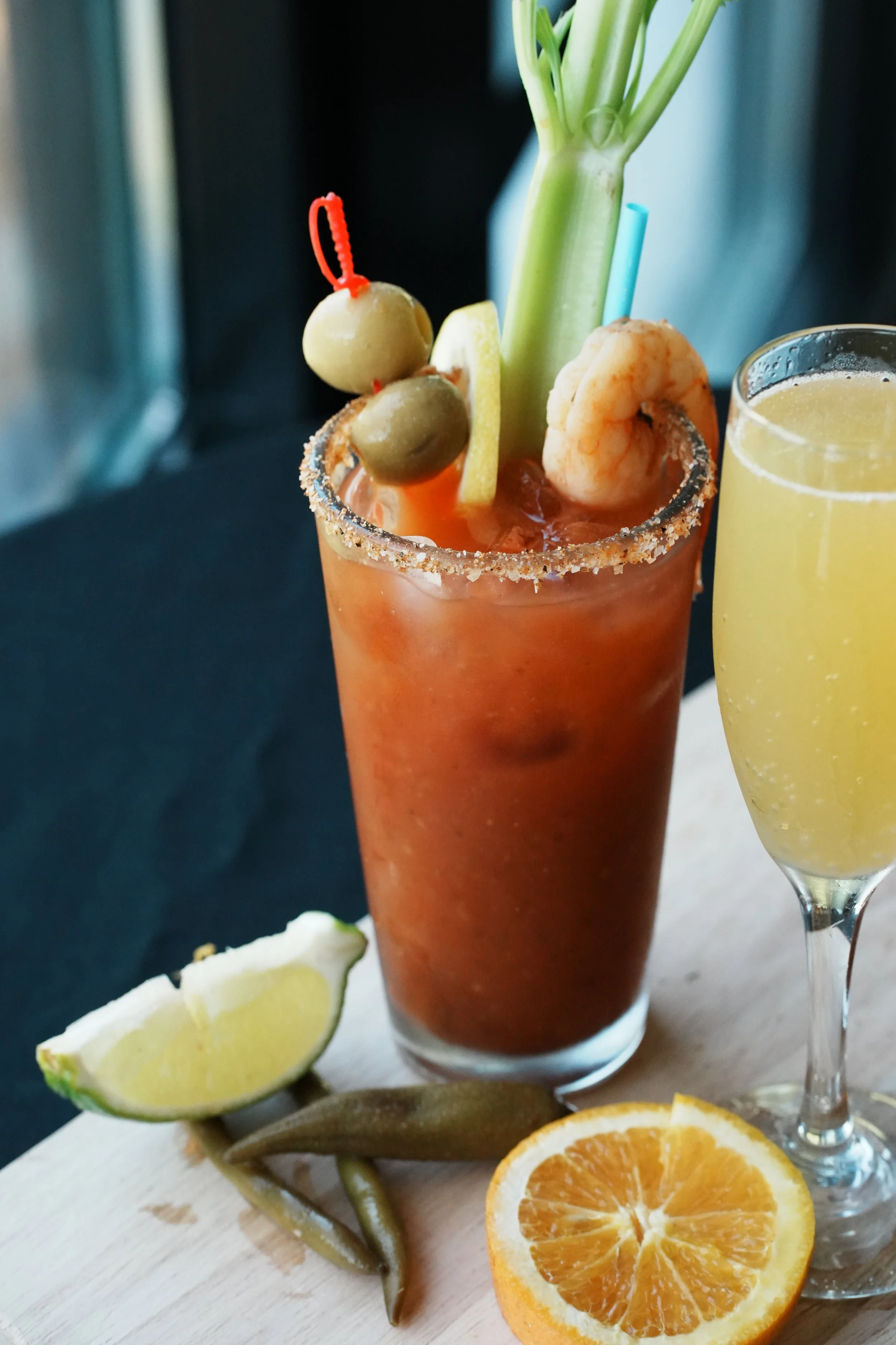 A Bloody Mary cocktail garnished with olives, lemon, celery, and shrimp, accompanied by a glass of mimosa with orange slices and a lemon wedge.