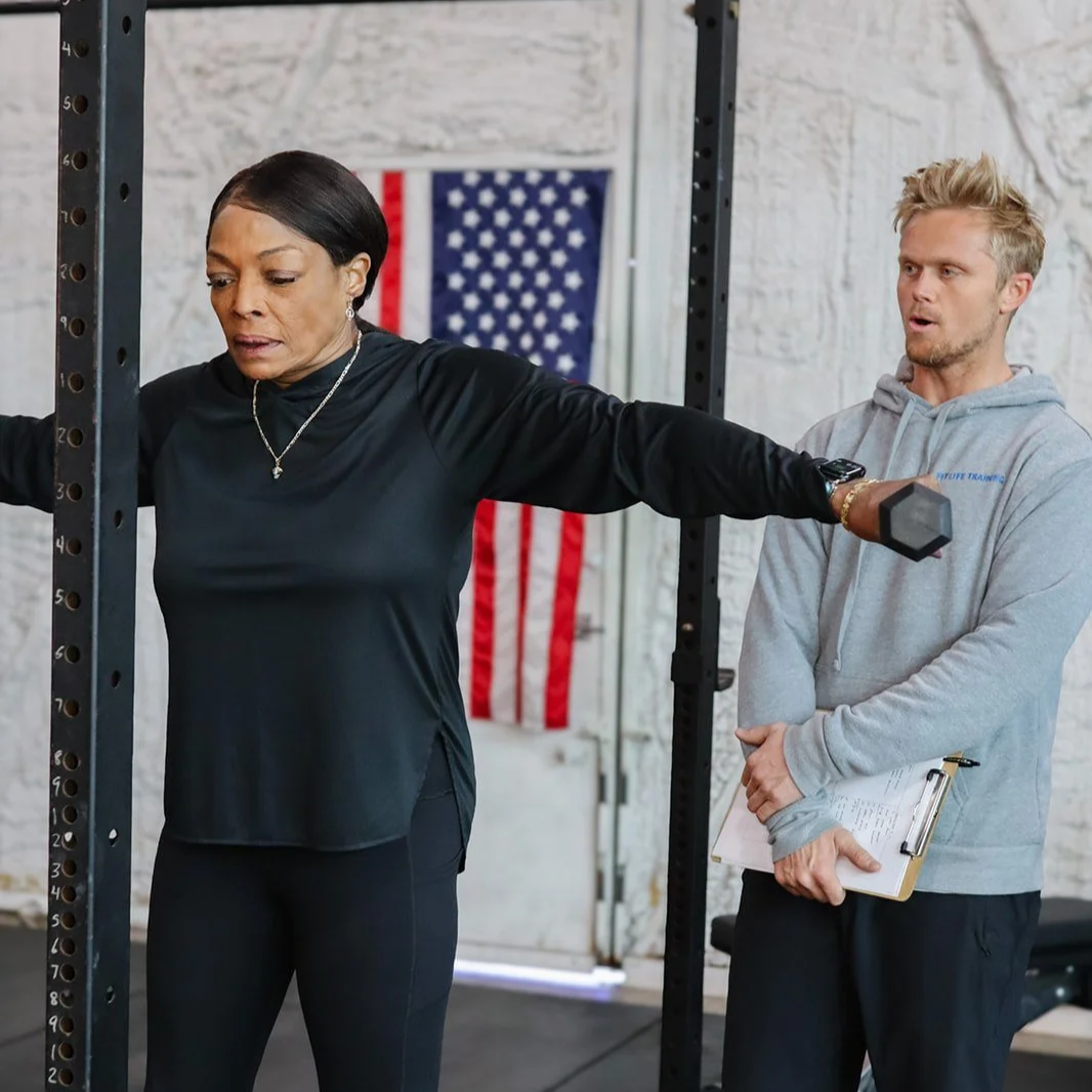 A man sitting on a barbell in a gym with a young girl standing on the barbell behind him.