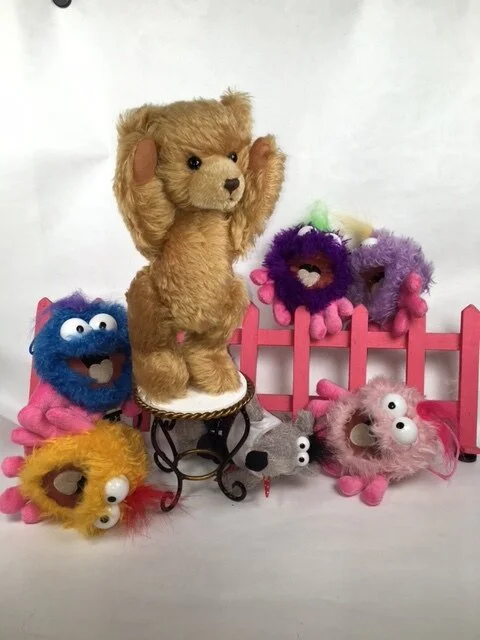 Verschiedene bunte Plüschtiere, darunter ein brauner Teddy auf einem kleinen Tisch, umgeben von bunten Wuscheltieren