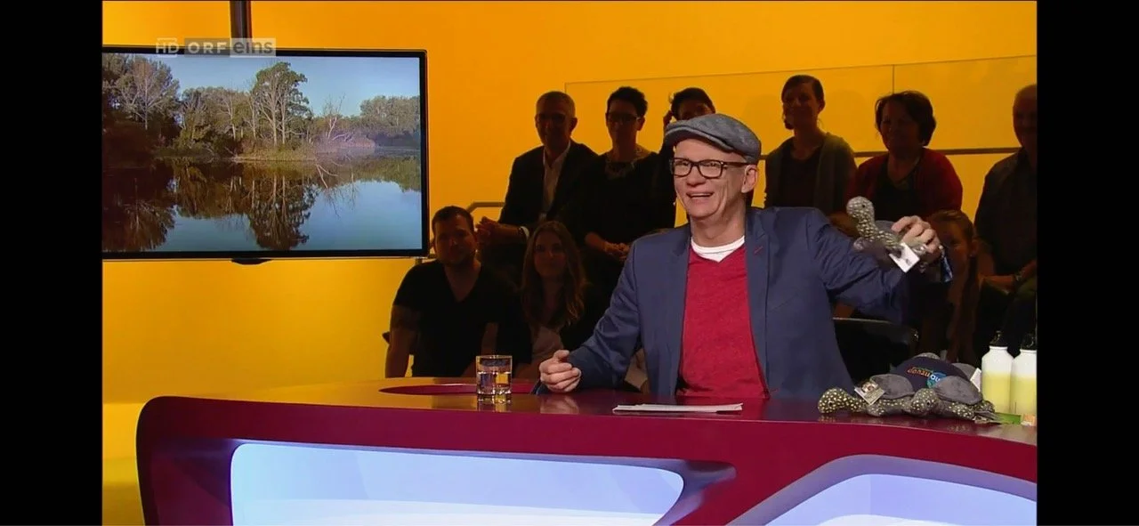 Ein Mann sitzt an einem rot-weißen Tisch in einem Studio, umgeben von mehreren Menschen, mit einem großen Bildschirm im Hintergrund, das eine Naturaufnahme zeigt.