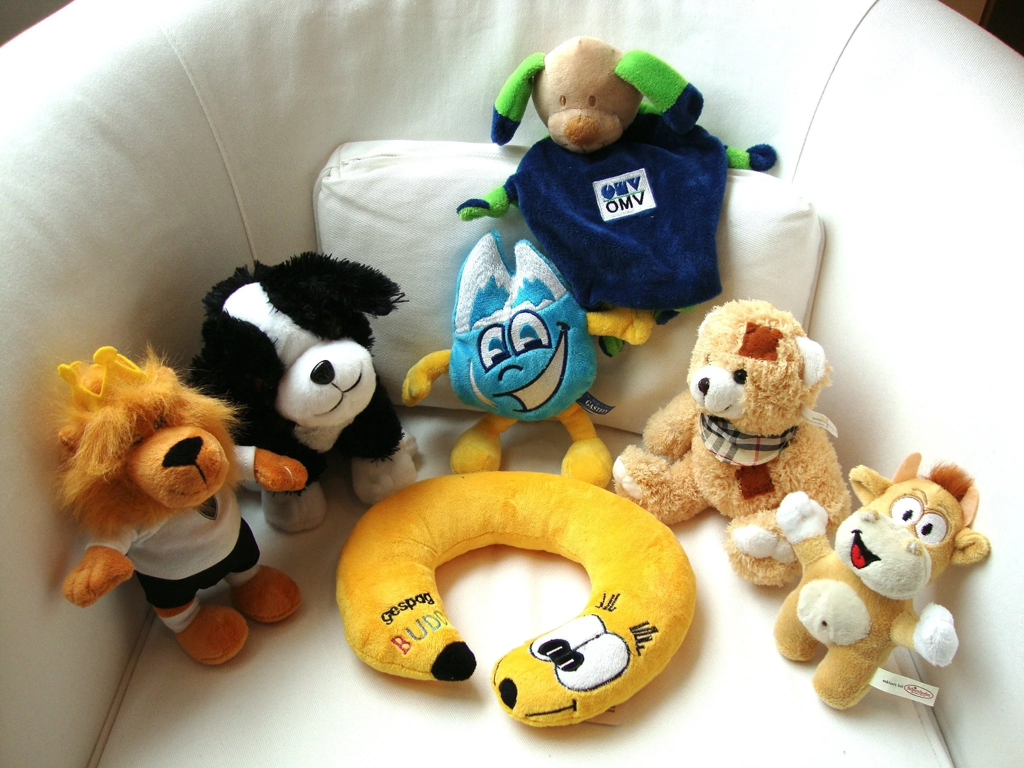 Verschiedene Spieltiere auf einem Sofa, darunter eine weiche Giraffe, ein schwarzer und weißer Hund, eine blauer Wasser-Tropfen mit lachender Gesichtsausdruck, ein brauner Löwe, ein brauner Affe, ein gelber Hase, und eine braune Steppenkatze.