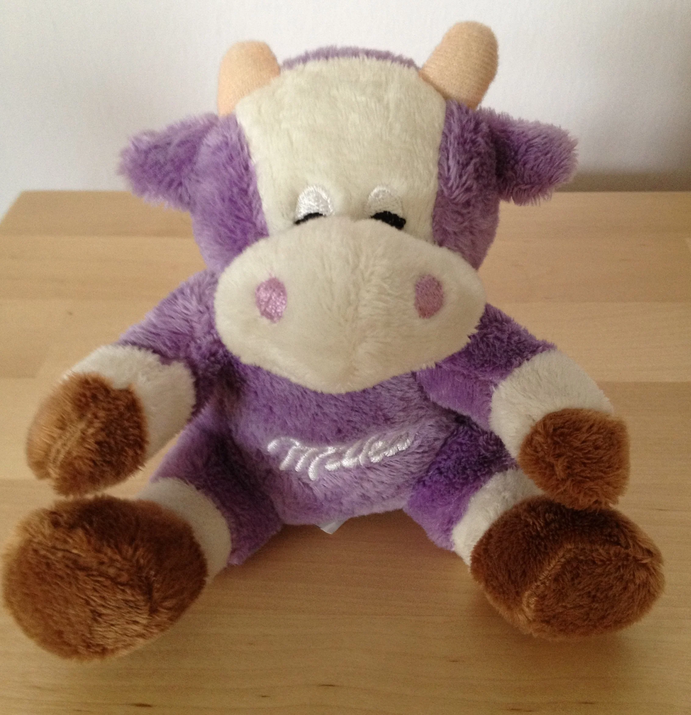 Ein Plüschtier in Form einer Kuh, das lila, cremefarbene und braune Farben hat, auf einem Holztisch sitzt.