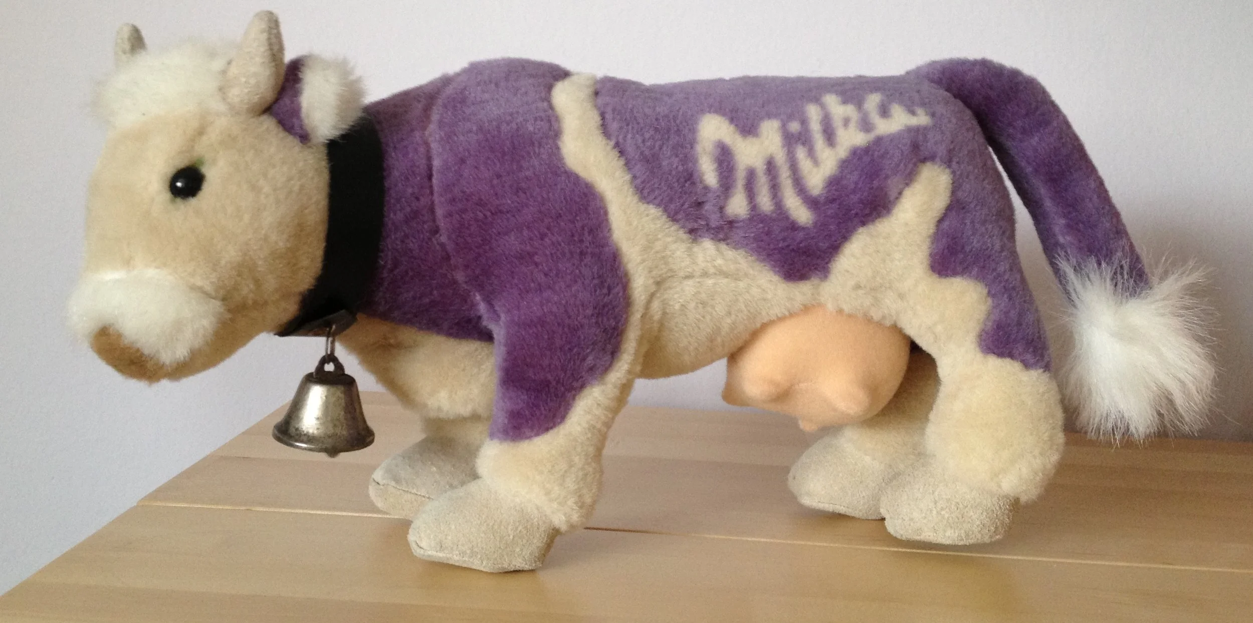 Plüschtier eines Hundes mit einer violetten Jacke, die das Wort 'Milka' trägt, und einer kleinen Glocke um den Hals, steht auf einem Holztablett